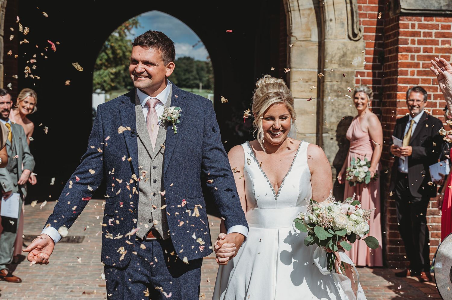 Happy newlyweds exit church ceremony as guests throw white confetti on sunny wedding day.