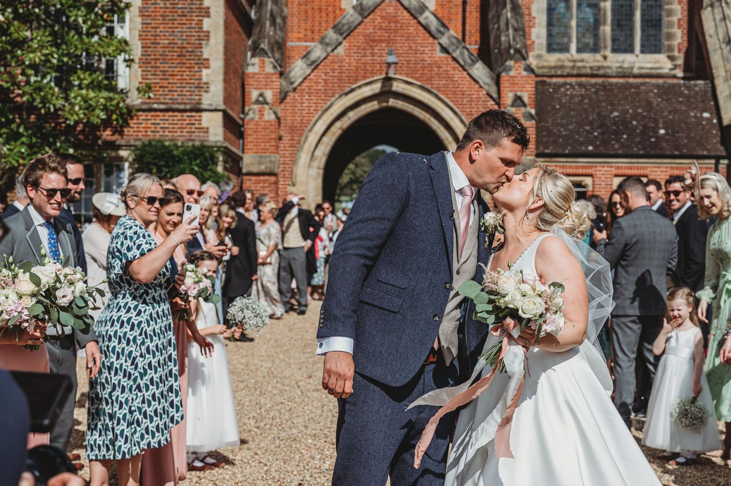 A newlywed couple shares a romantic moment outside a historic brick church while wedding guests look on and celebrate.