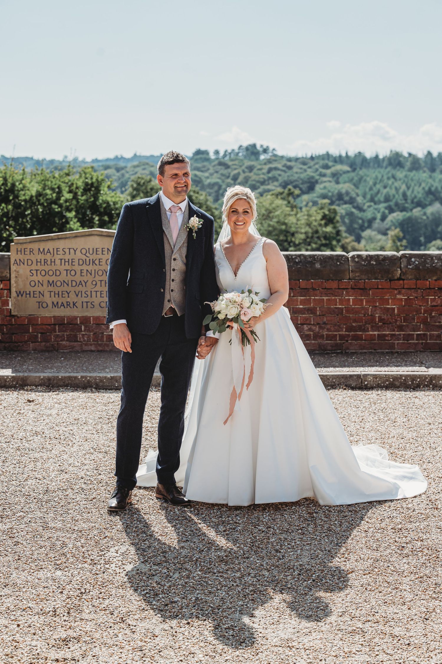 A bride and groom stand together outdoors on a sunny day against a brick wall with scenic countryside views.