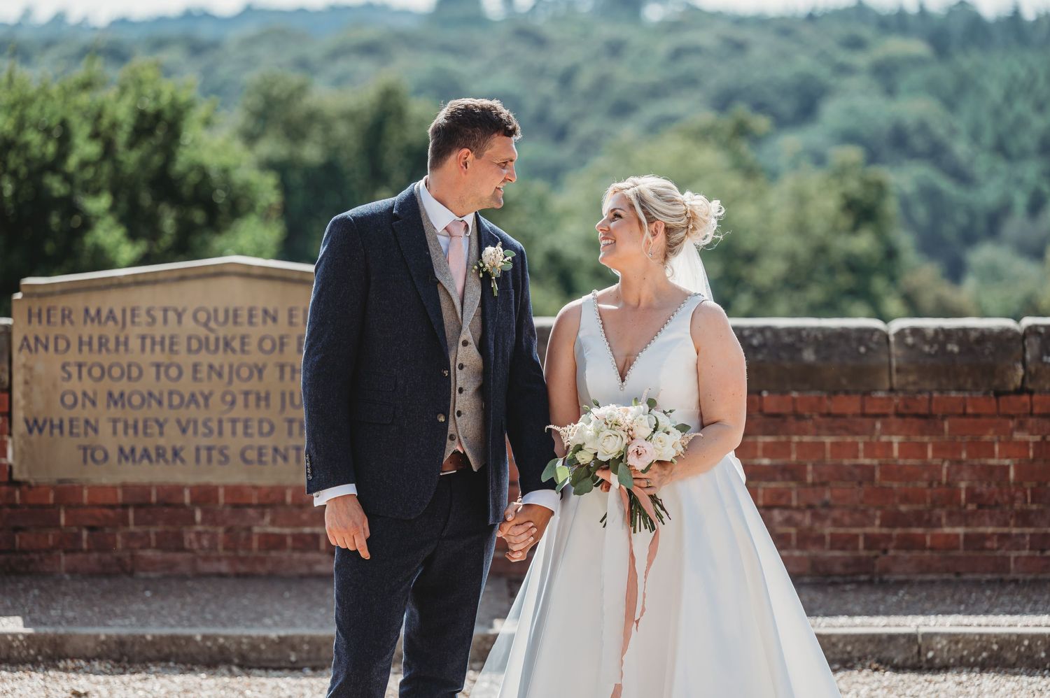 Wedding couple walk along historic brick wall on sunny day holding bouquet of flowers.
