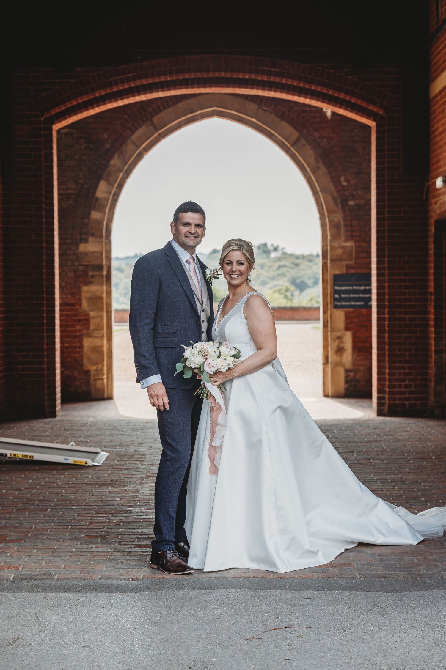 A bride and groom pose under a red brick archway, with the bride holding a white bouquet.