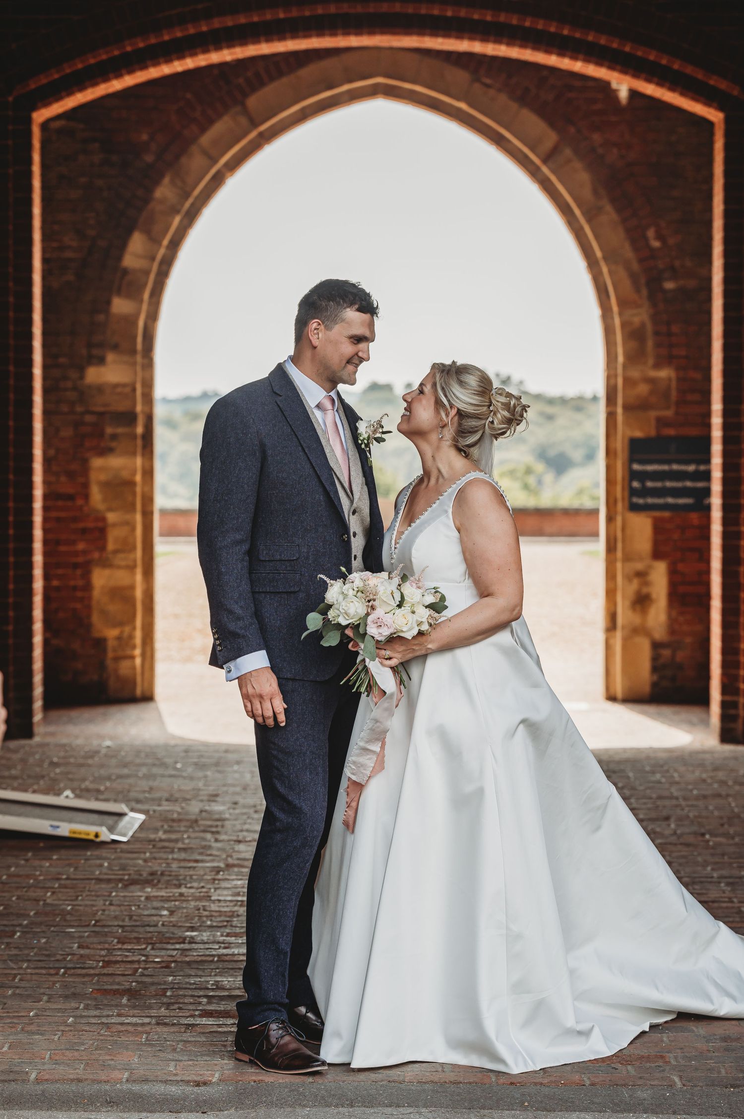 A romantic wedding couple standing under a brick archway, the bride in white holding a bouquet and groom in navy suit.