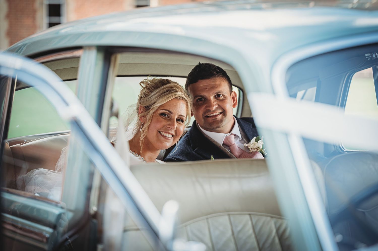 A couple sits in a classic vintage car, smiling through the window on their wedding day.