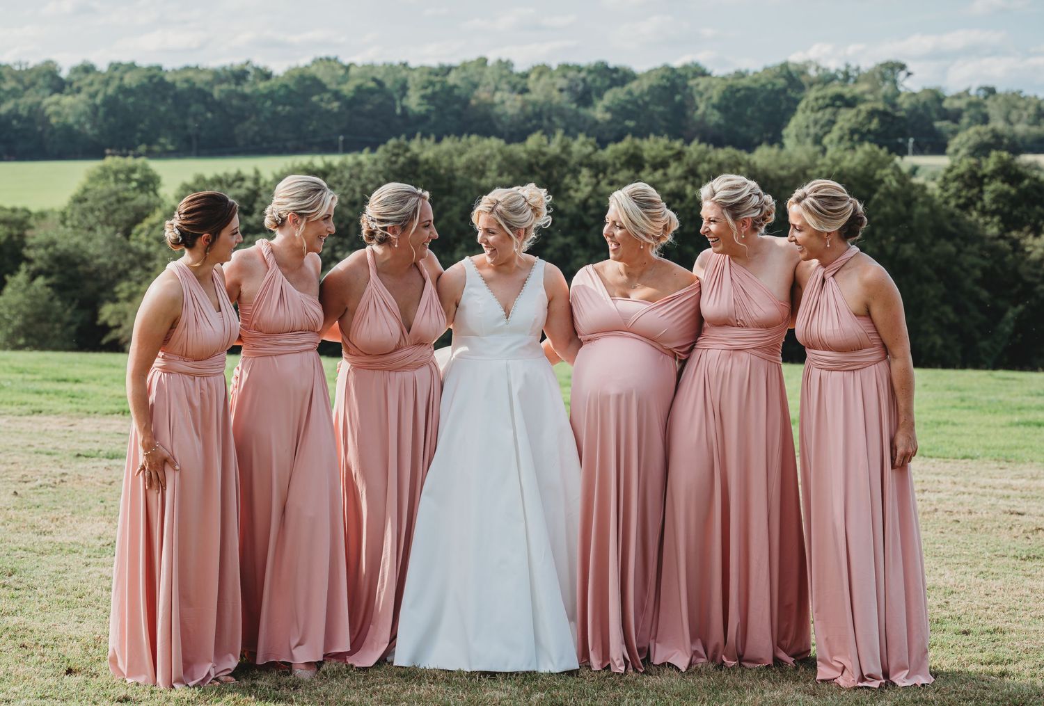 Seven women in matching dusty rose bridesmaid dresses gather outdoors with a bride in white against a scenic countryside view.