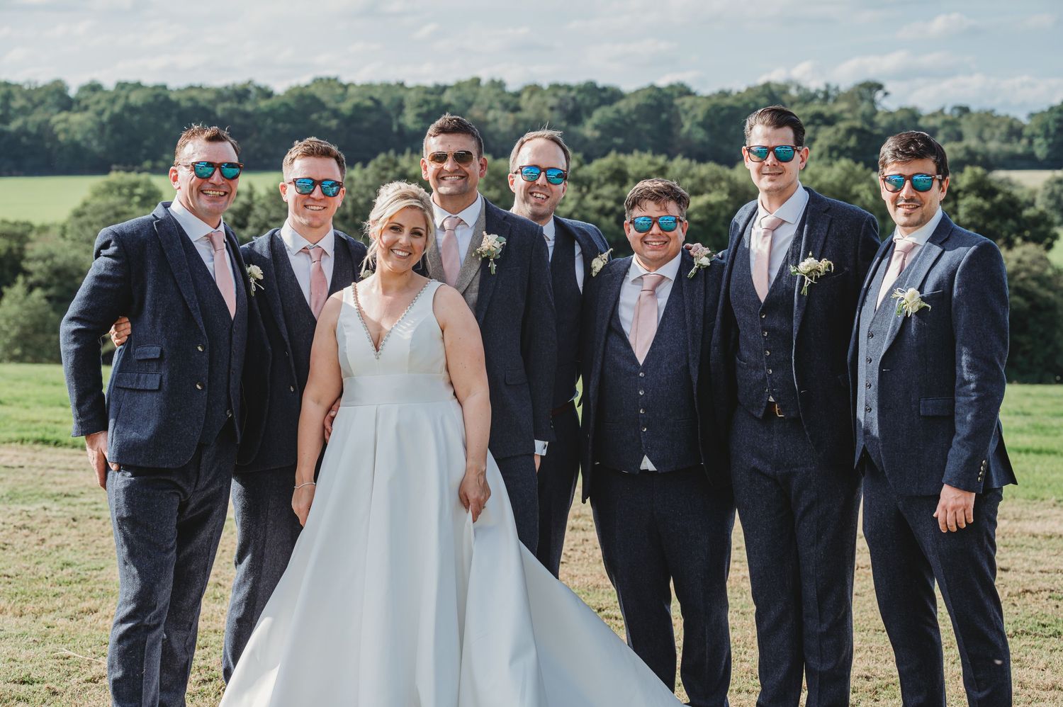 Wedding party wearing navy suits and sunglasses pose with a person in white dress in front of scenic countryside view.
