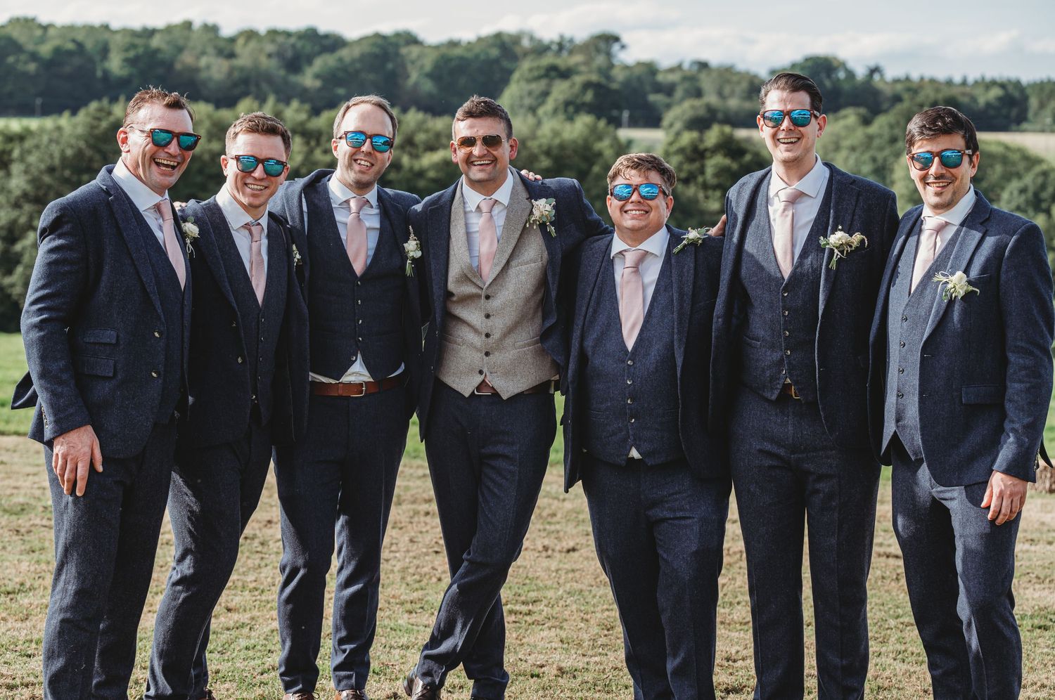A group of seven groomsmen in navy blue suits and sunglasses pose together outdoors on a sunny hillside.