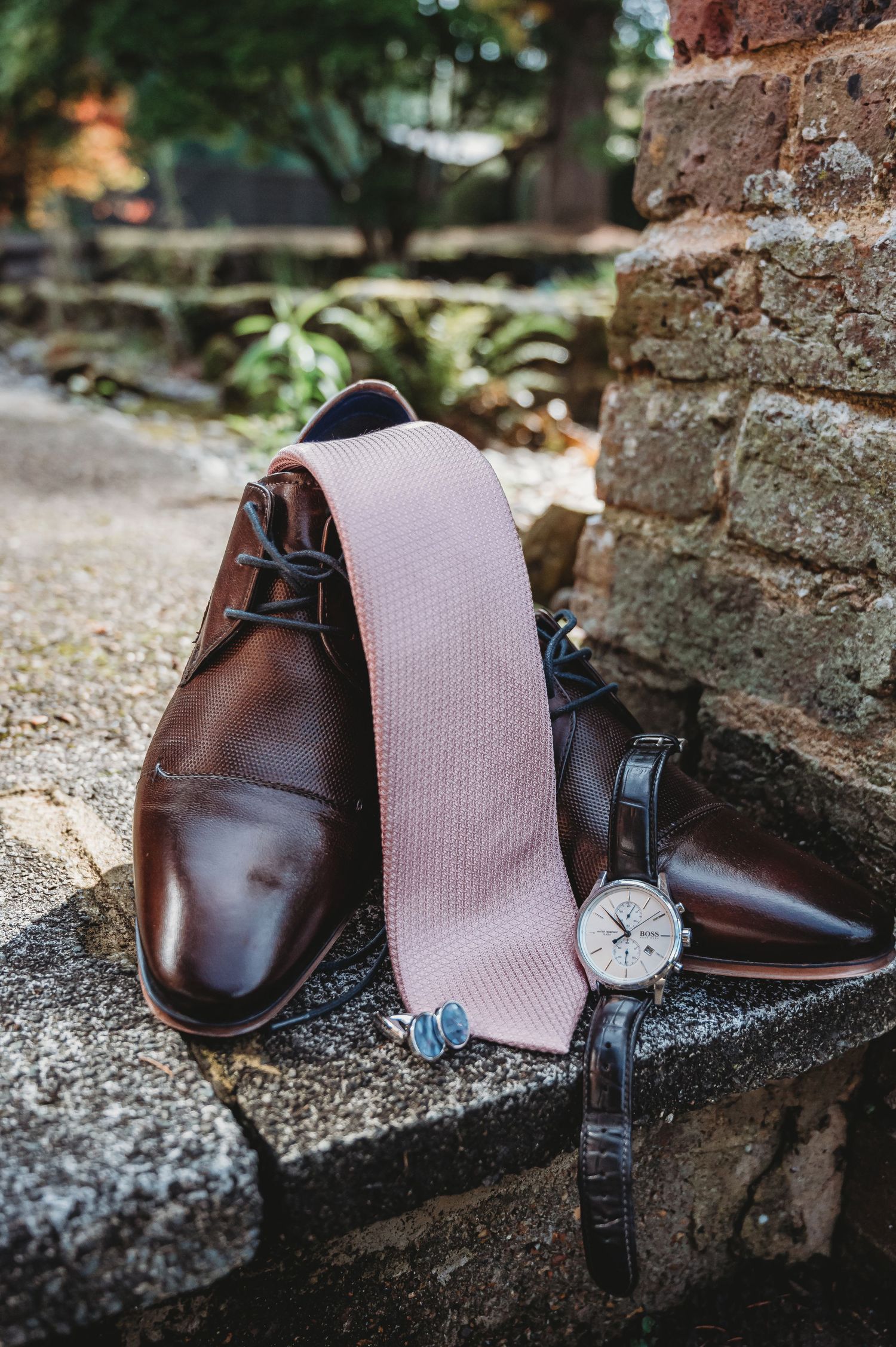 Brown leather dress shoes arranged with a blush pink tie and silver watch on stone steps near a brick wall.