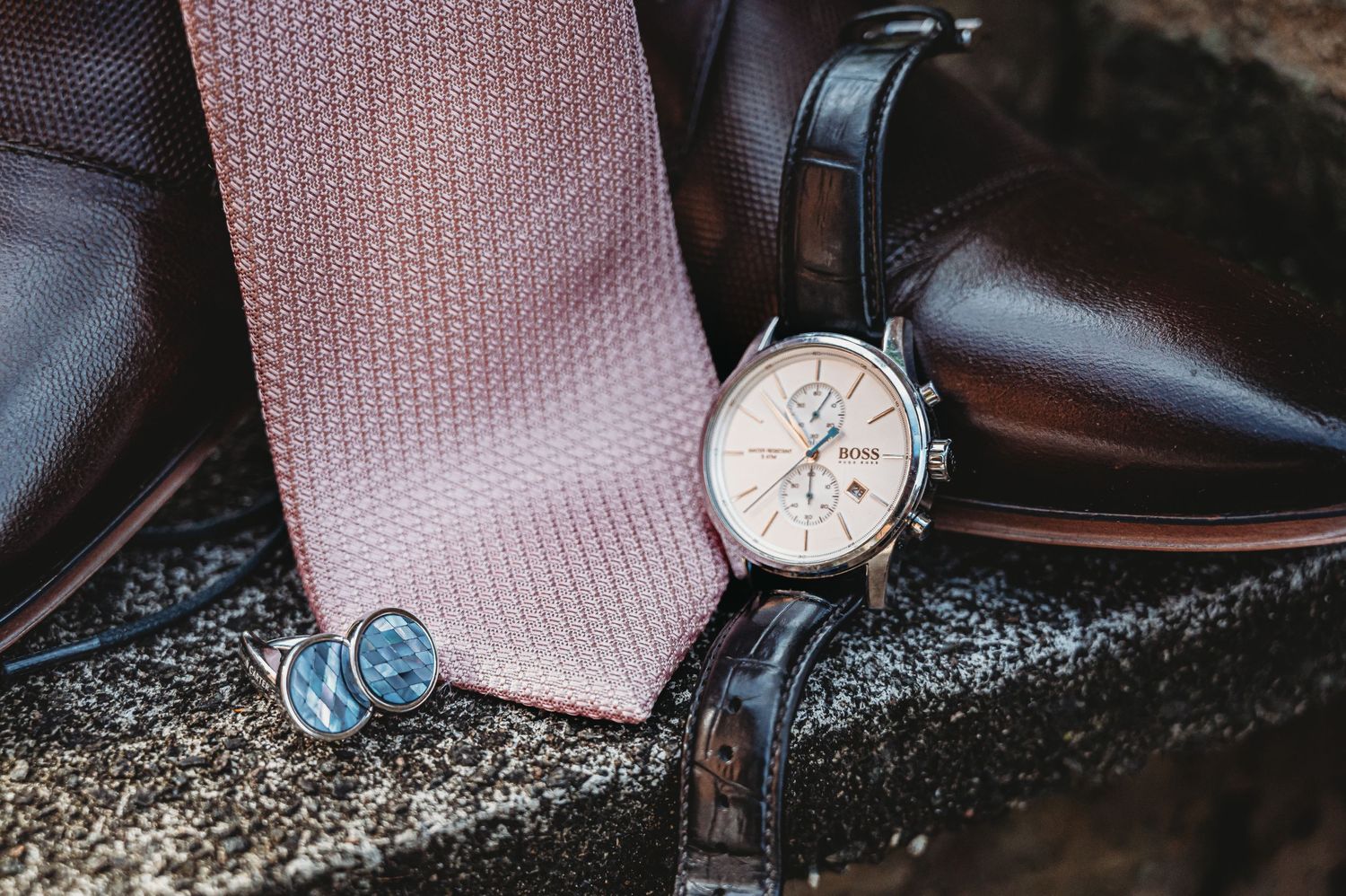 A sophisticated mens accessory flatlay with a pink silk tie, leather dress shoes, and elegant chronograph watch.