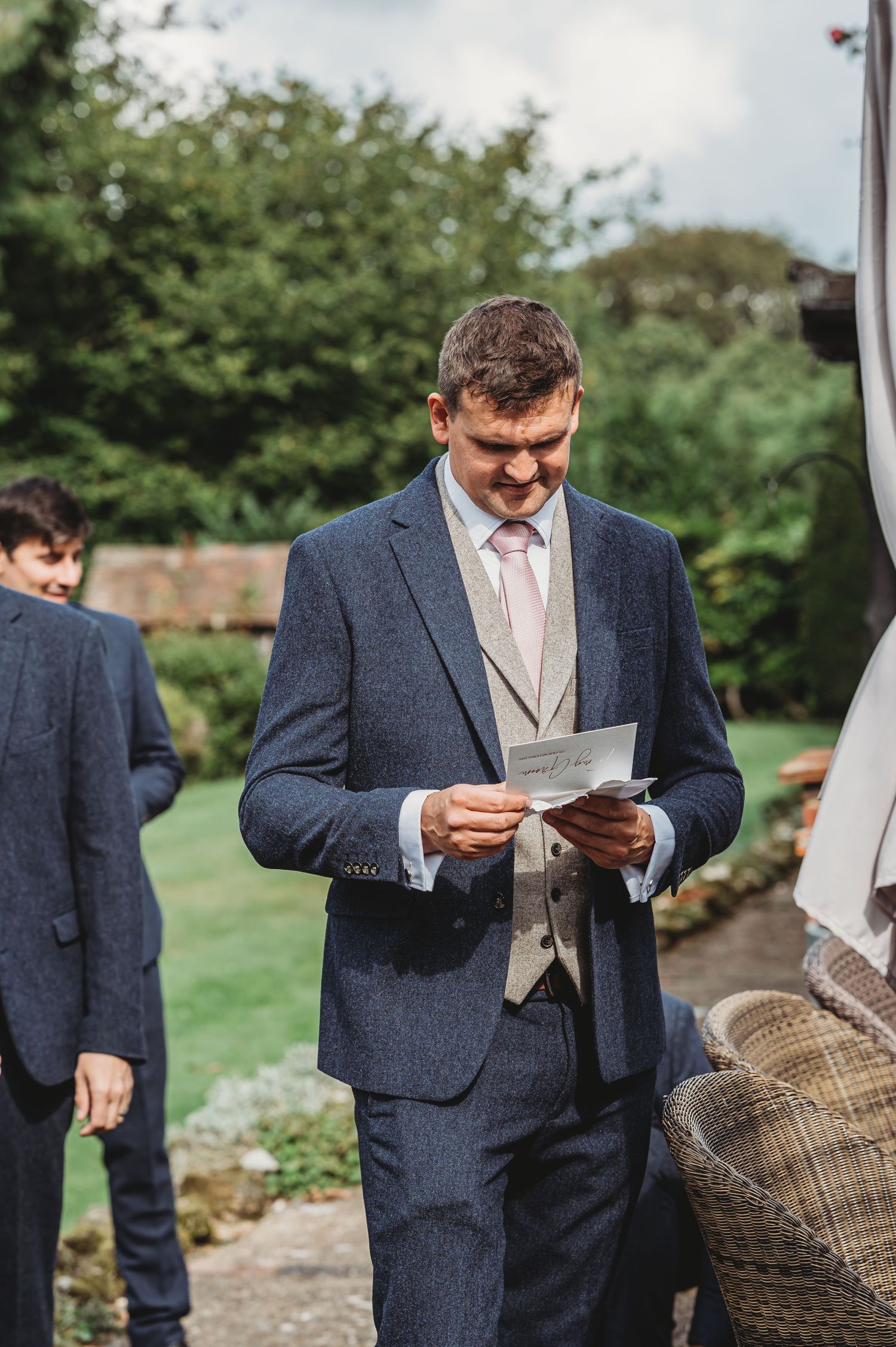 A person in a navy suit and grey waistcoat reads from papers while standing outdoors at a formal event.