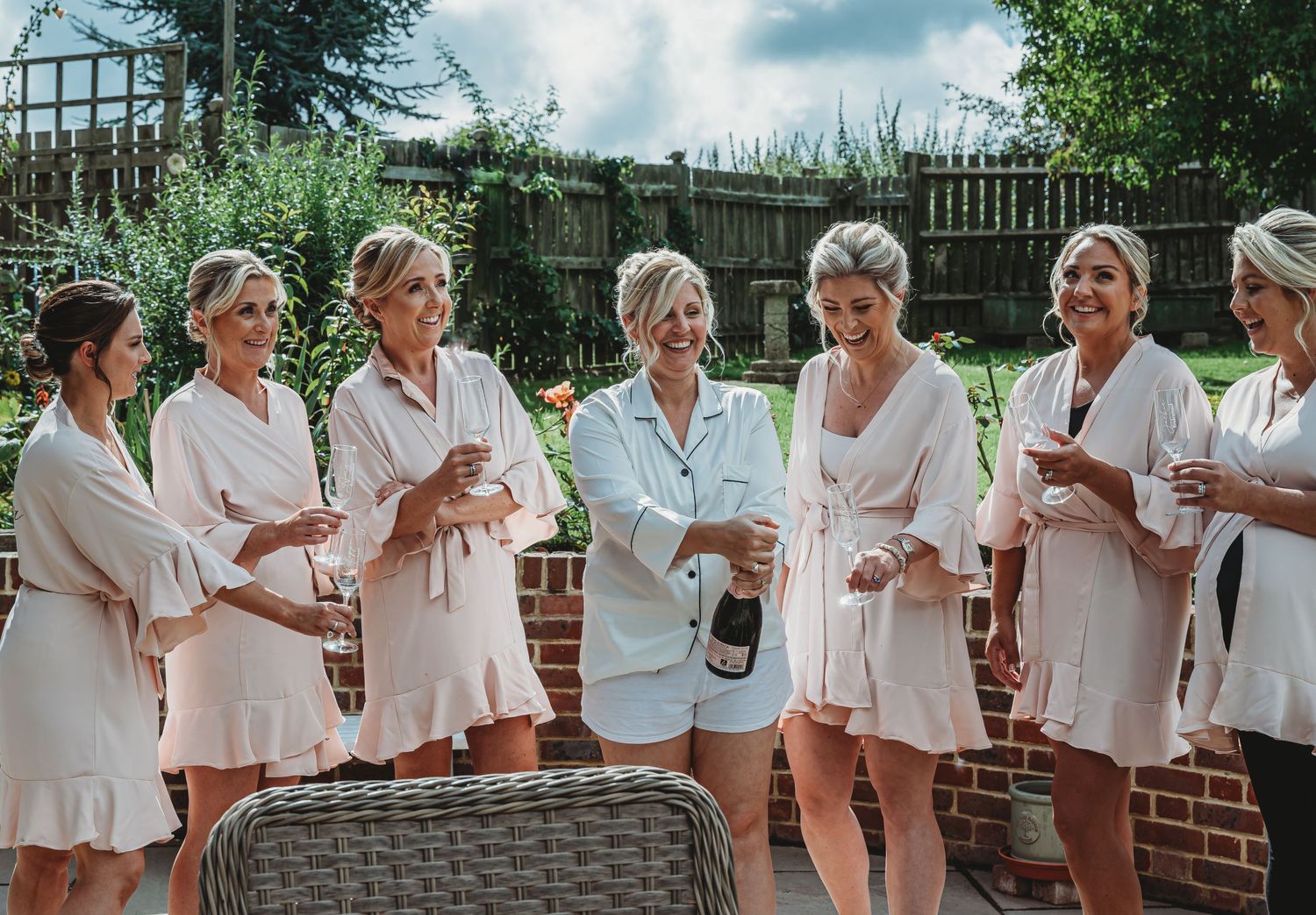 A group of people in matching white and blush-colored robes gather for a celebratory pre-wedding moment outdoors.