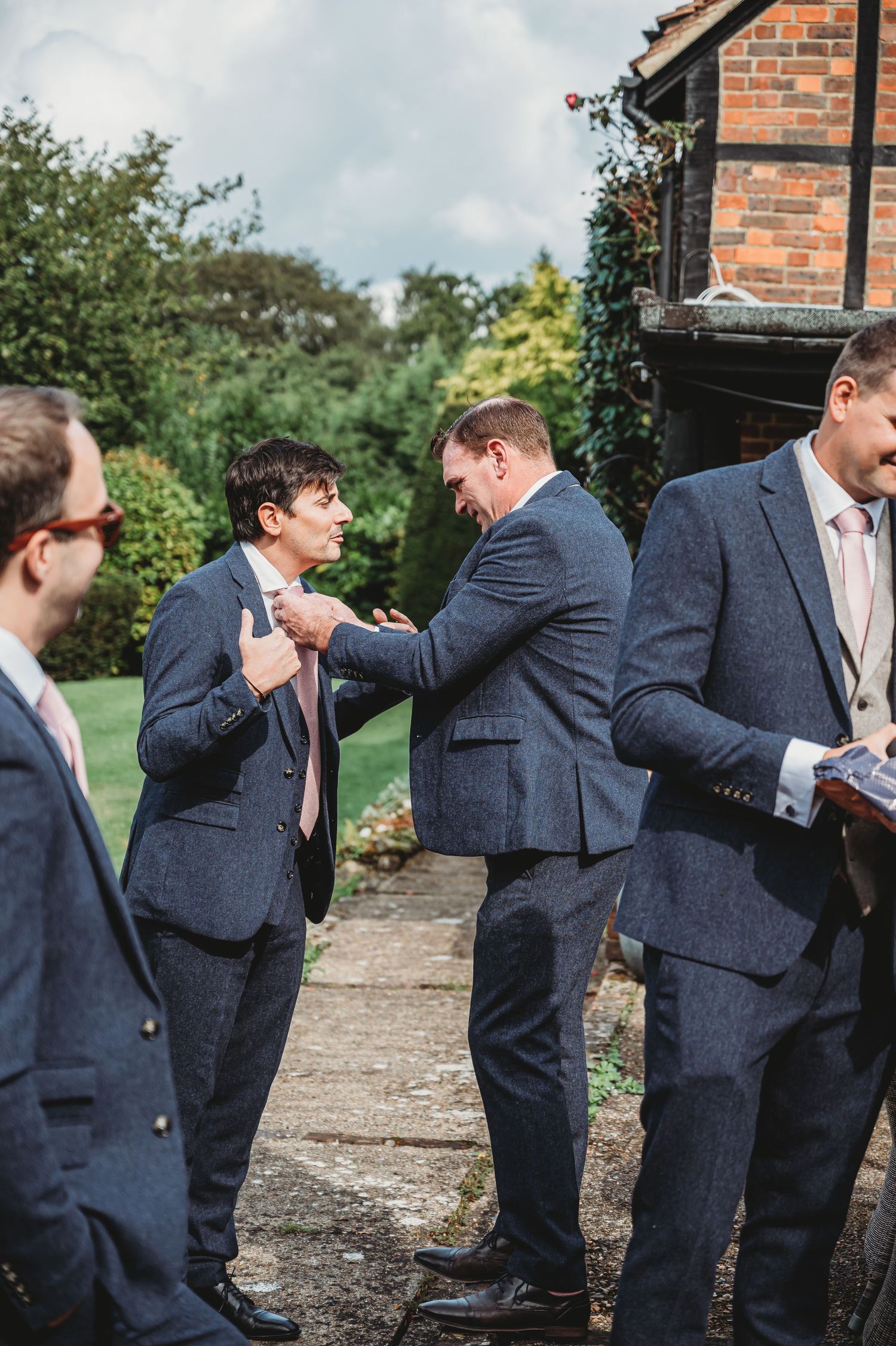 Groomsmen in navy suits help adjust each other's ties and boutonnières before a wedding ceremony outdoors.