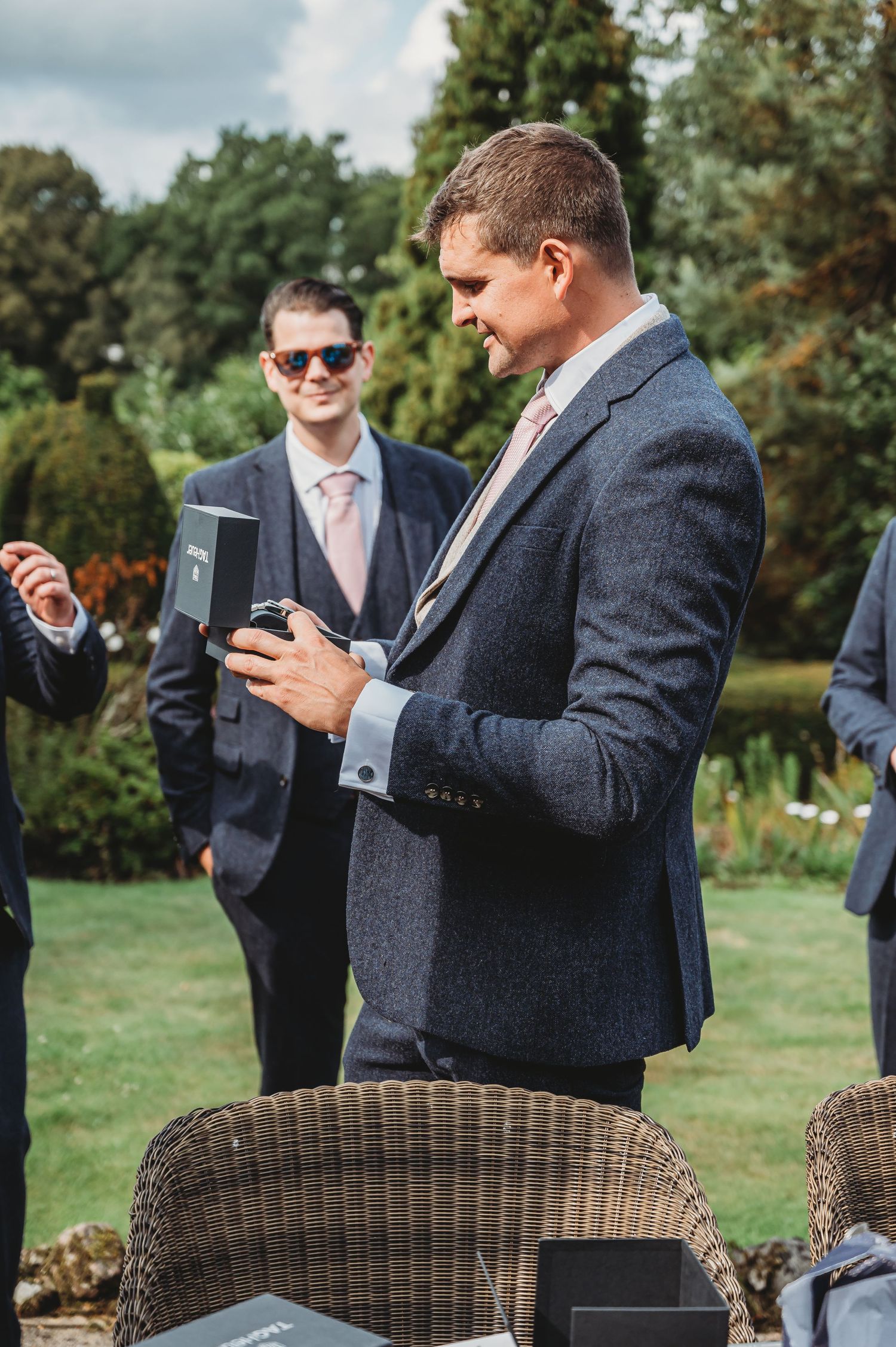 Attendees in grey suits checking their phones at an outdoor gathering with wicker furniture.