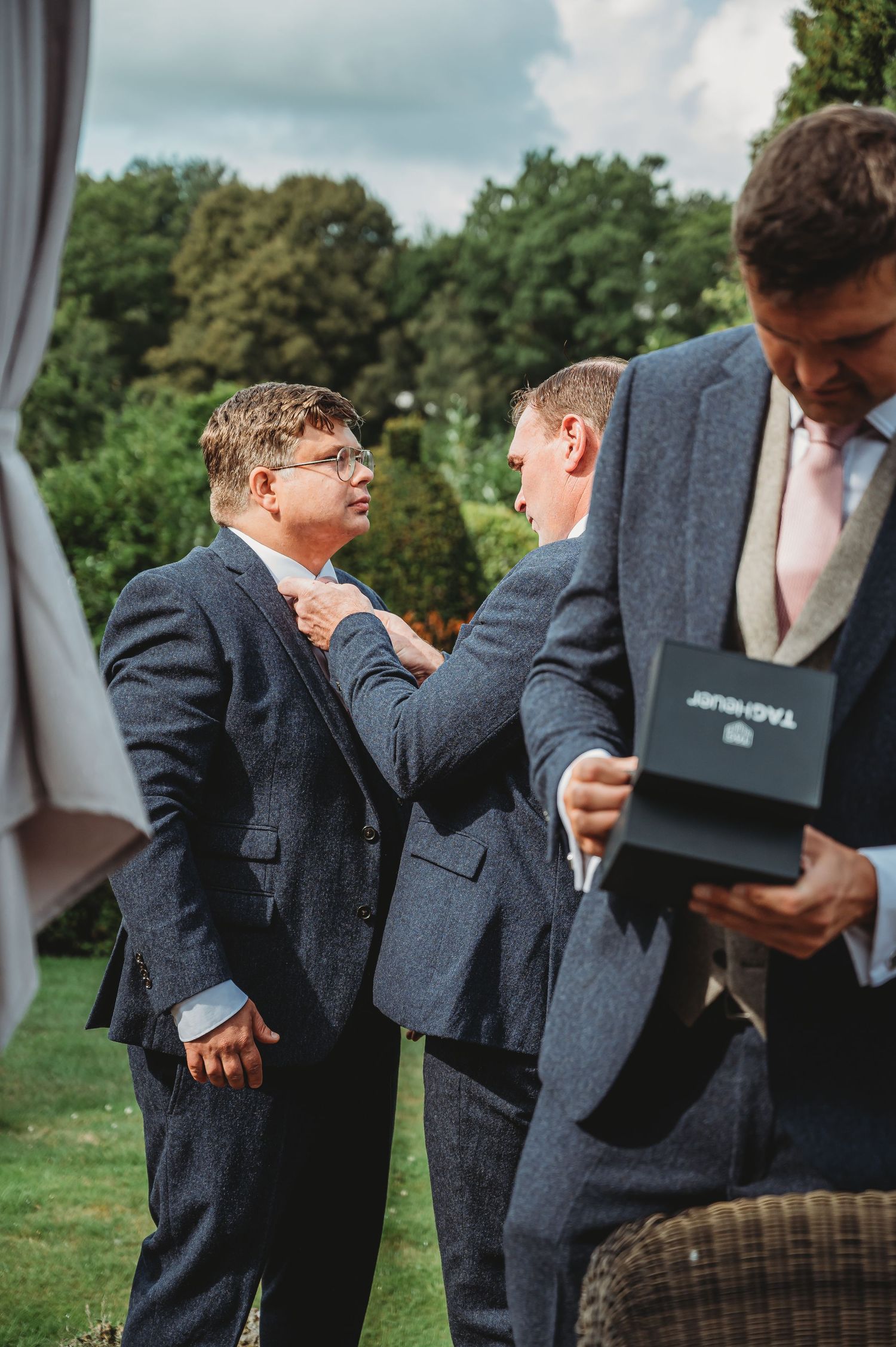 A group adjusting formal attire and checking a small box during an outdoor event on a cloudy day.