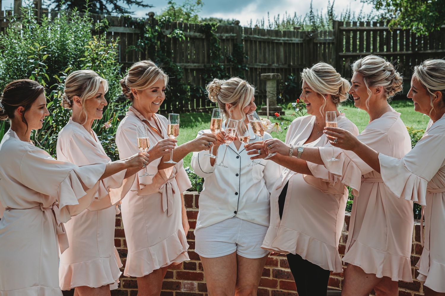 A group in matching pink robes toasting with champagne glasses in a sunny backyard setting.