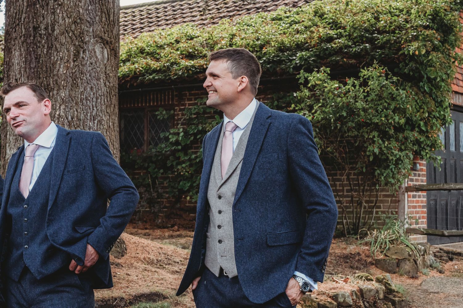 Two groomsmen in navy suits and light grey waistcoats stand casually outside a brick building with greenery.