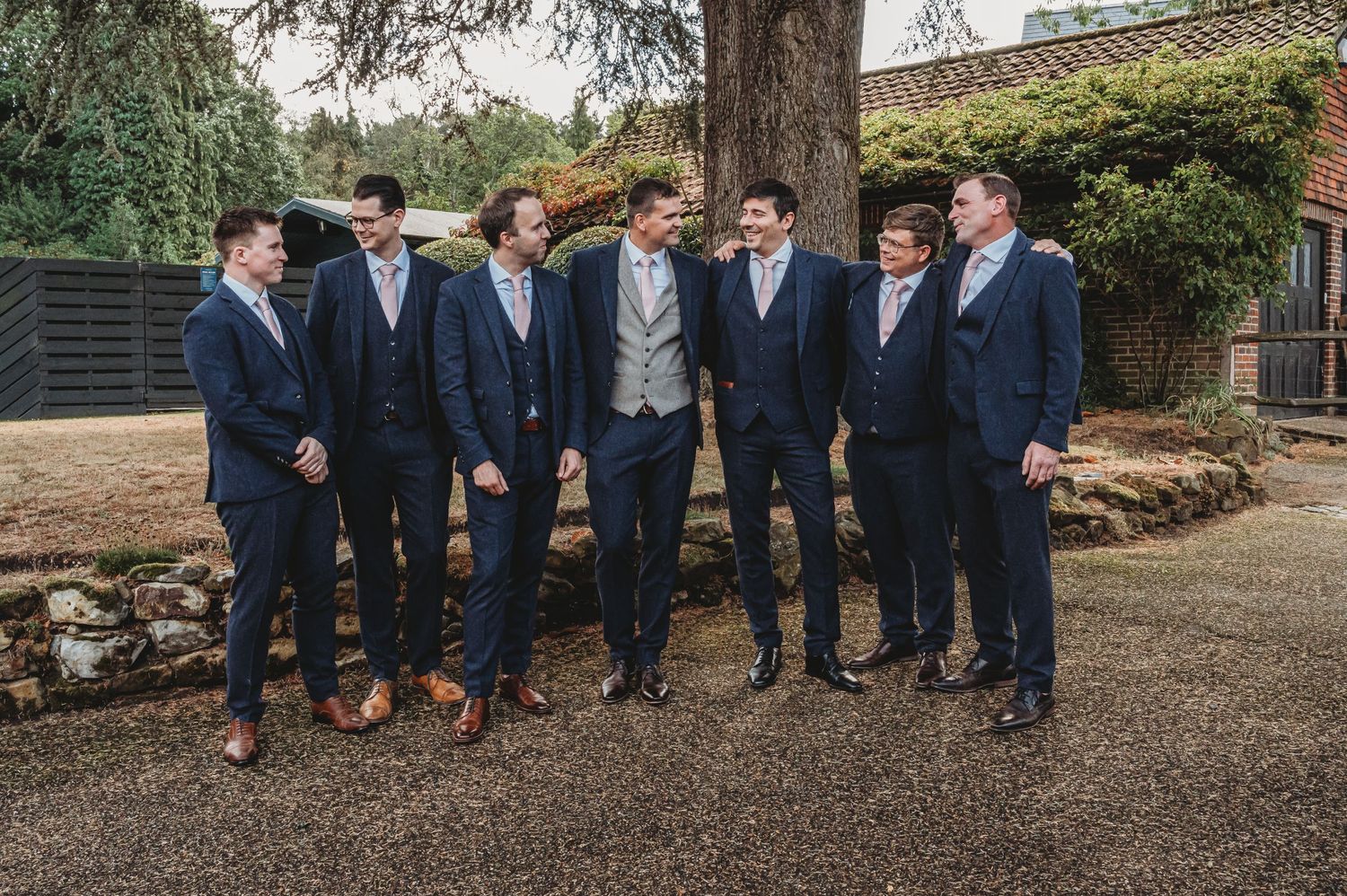 A group of seven men stand together outdoors wearing coordinated navy suits and formal attire for a wedding occasion.