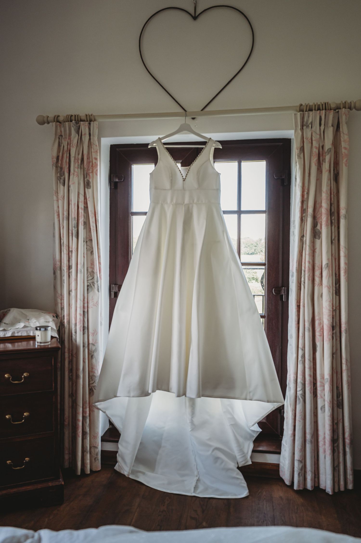 A white wedding dress hangs elegantly against French doors with floral curtains, beneath a metal heart wall decoration.