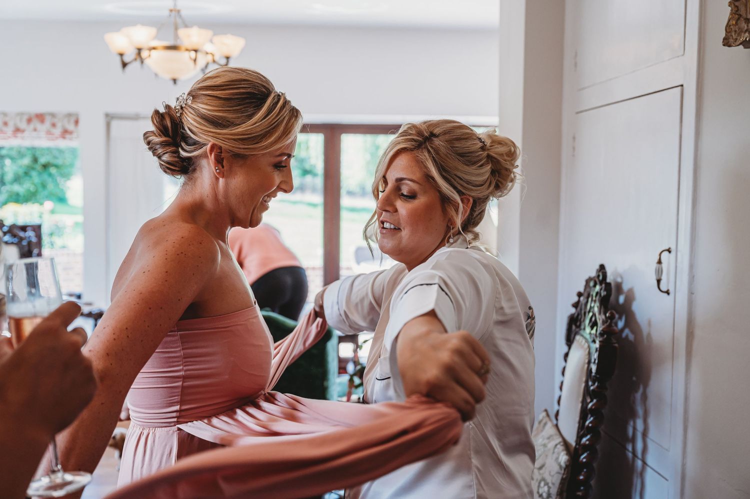 Two people share a joyful moment while getting ready in a bright room with a chandelier visible in the background.