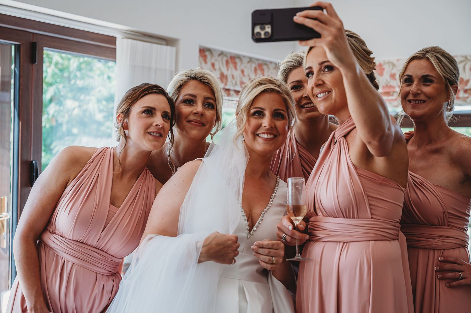 A group in matching pink bridesmaid dresses gather around for a celebratory selfie with the bride in white.