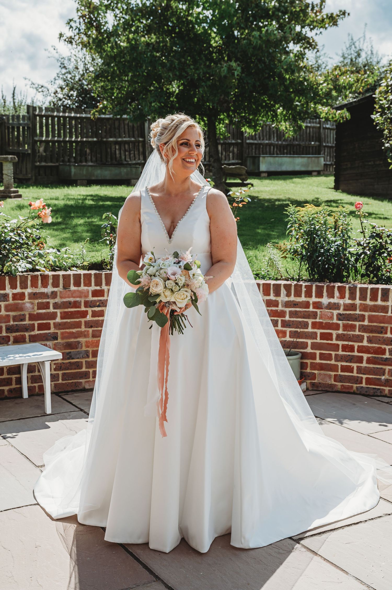 A bride in a flowing white v-neck wedding gown holds a blush and white bouquet in a garden setting with brick walls.
