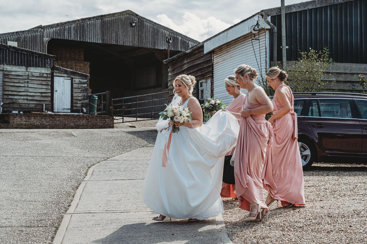 Several people in formal wedding attire walk together outside rustic barn buildings on a sunny day.