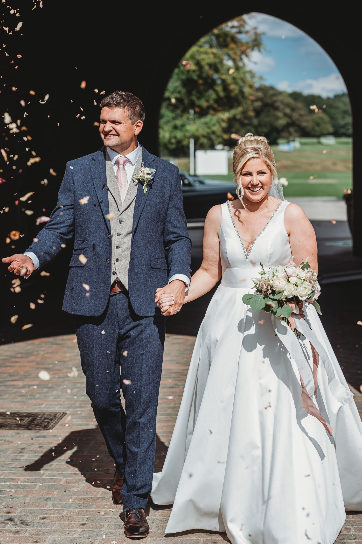 A bride in a white gown and groom in navy suit walk together on a sunny day while confetti falls around them.