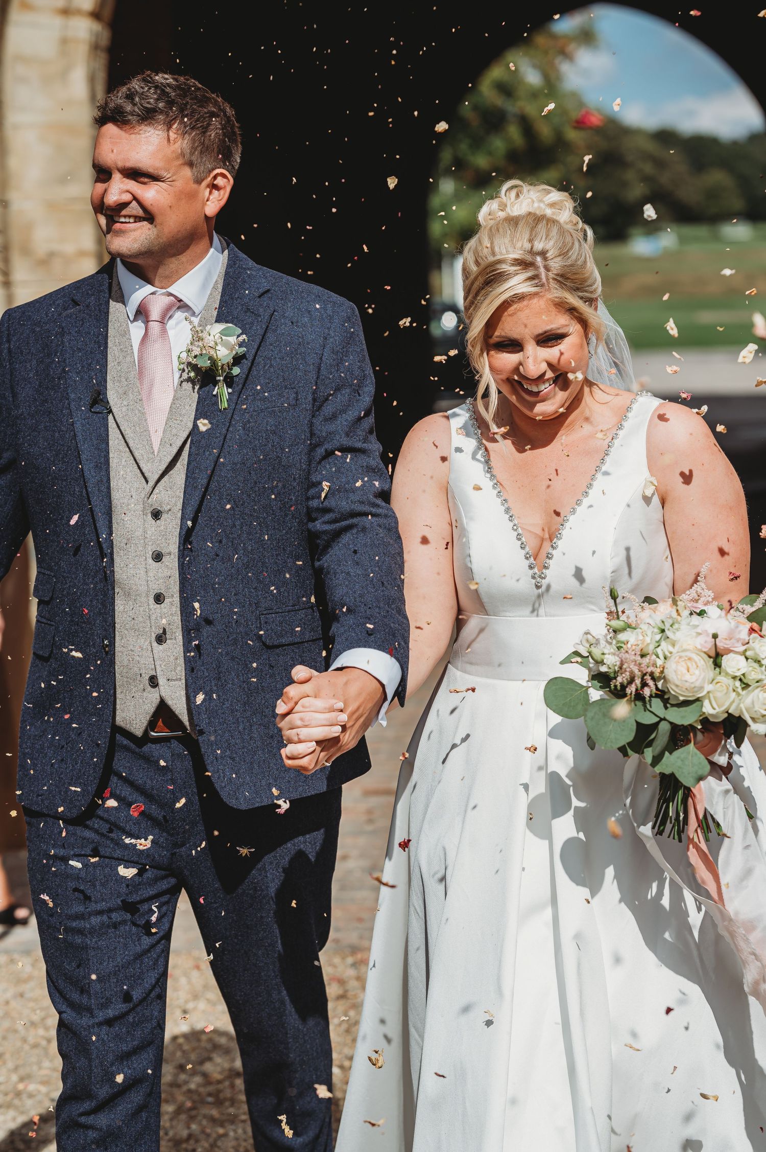 Newlywed couple walking together in formal attire while confetti falls around them on sunny wedding day.