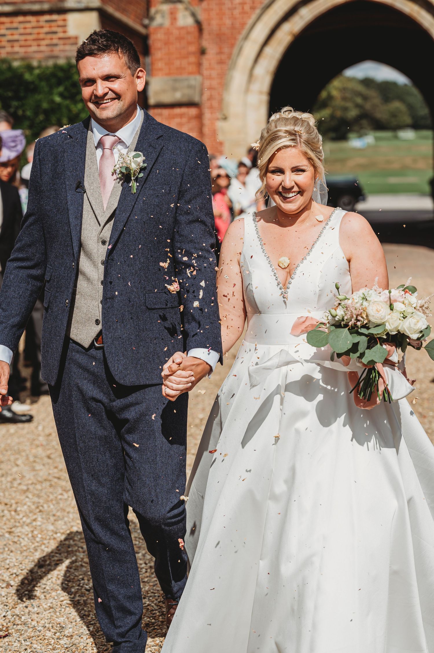 A joyful wedding couple walks together while confetti falls around them outside a brick building with an arched doorway.