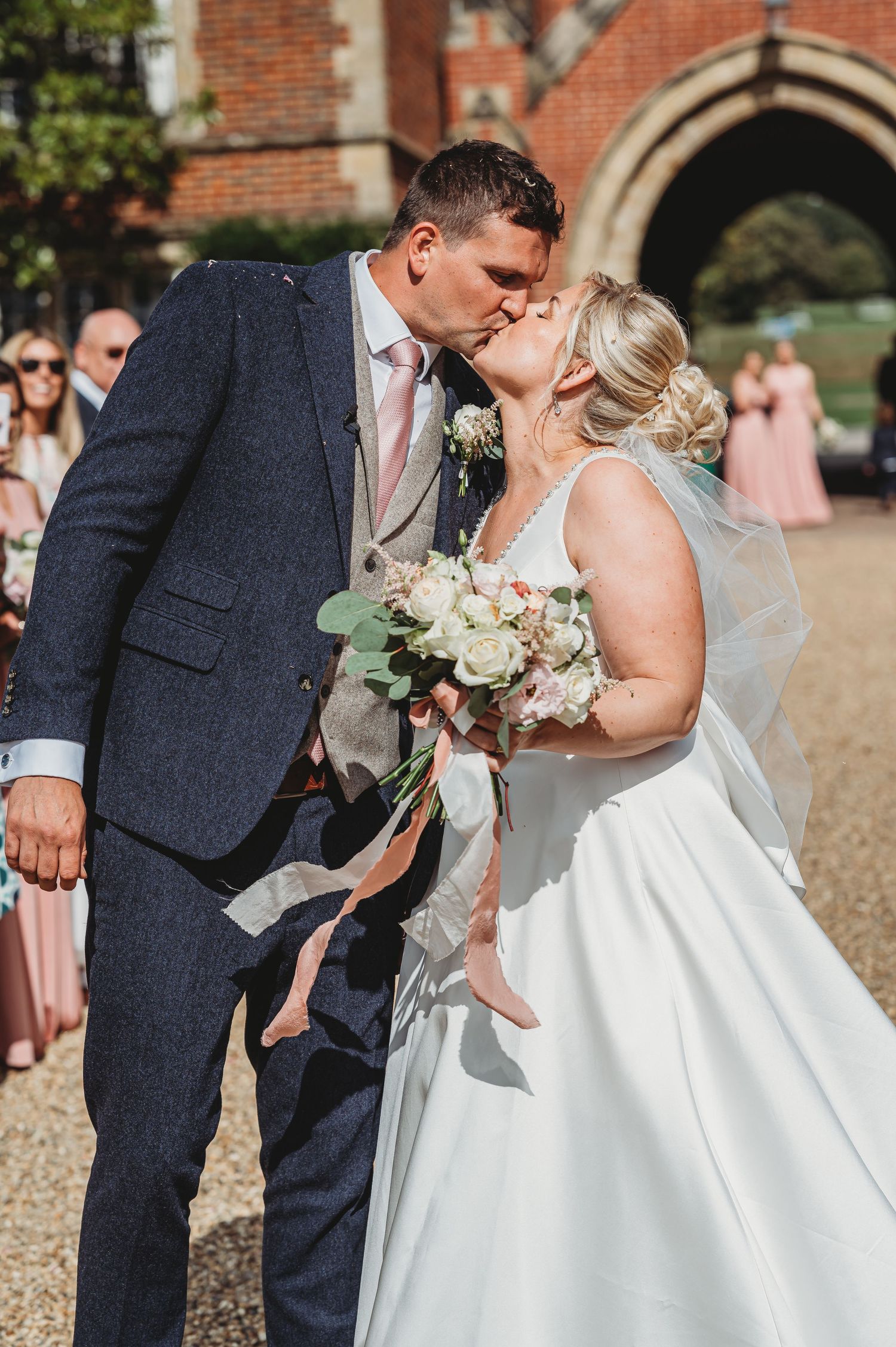 A joyful wedding couple shares a tender moment outside a brick church while holding a soft pastel bouquet.
