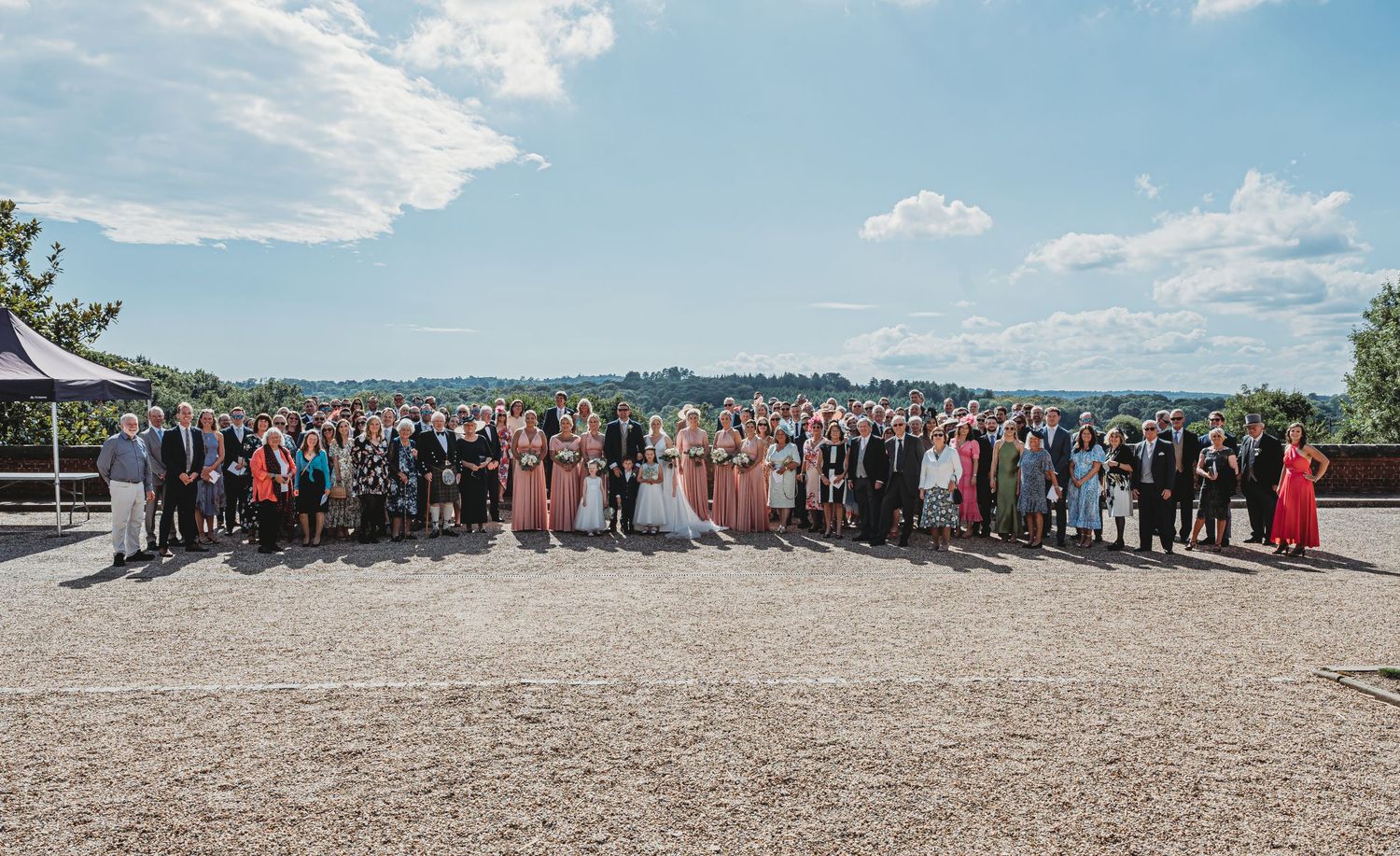 Large group of wedding guests gathered outdoors on a terrace overlooking scenic landscape on a sunny summer day.