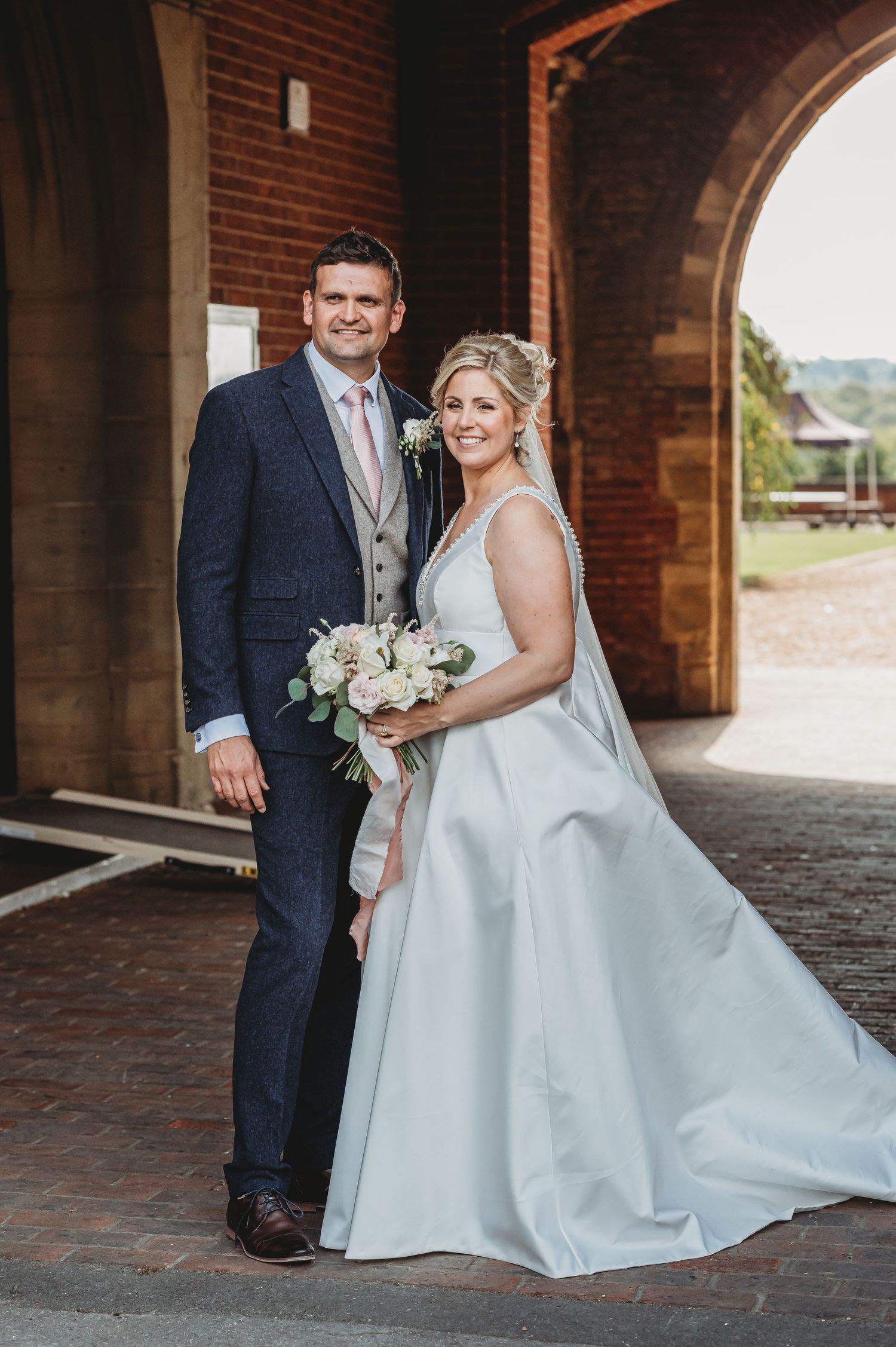 A newlywed couple poses under an archway at a brick venue, with the bride wearing a flowing white gown and holding flowers.