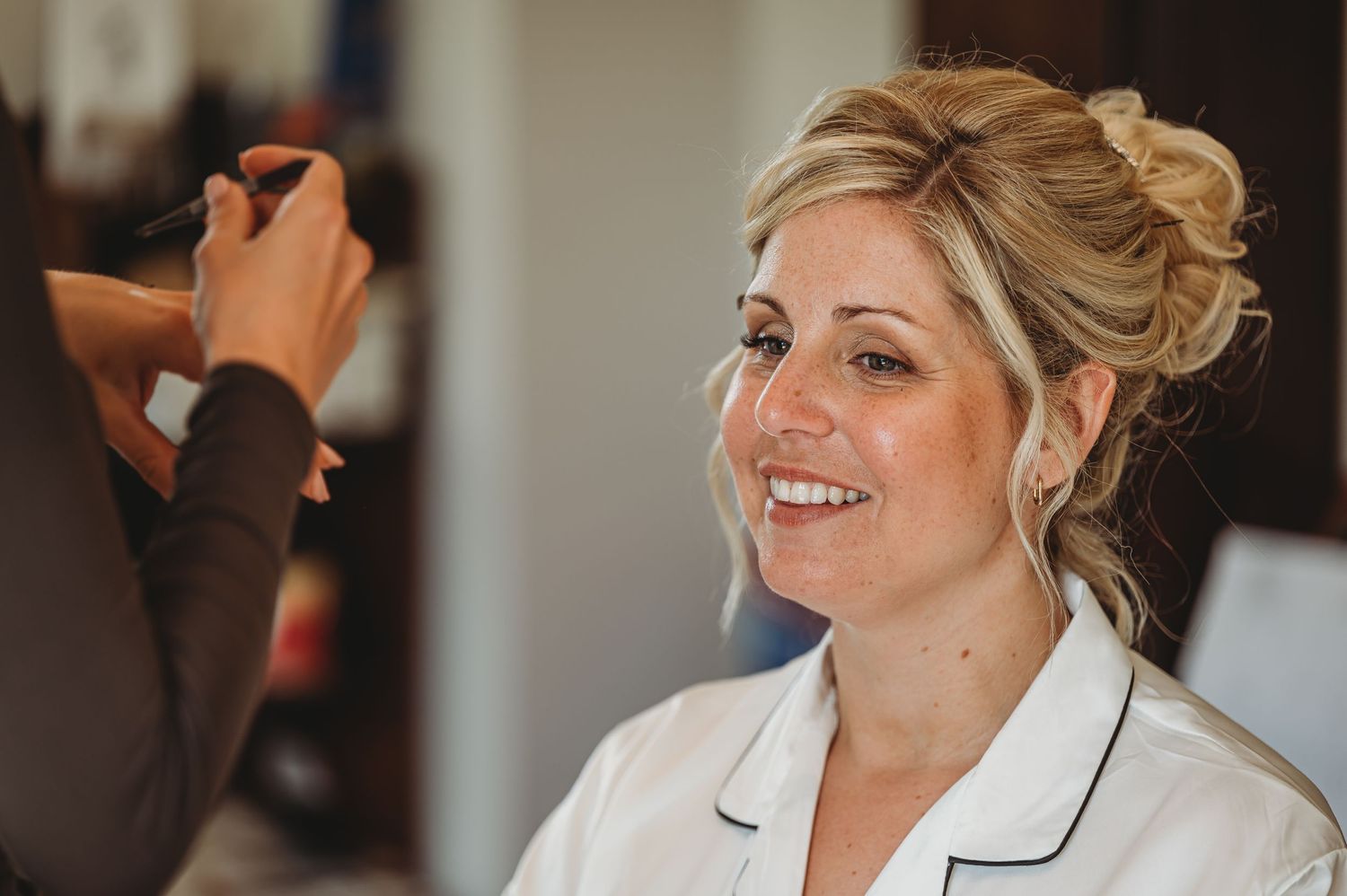 Getting ready for the big day, a bride in white robe smiles while having her makeup applied.