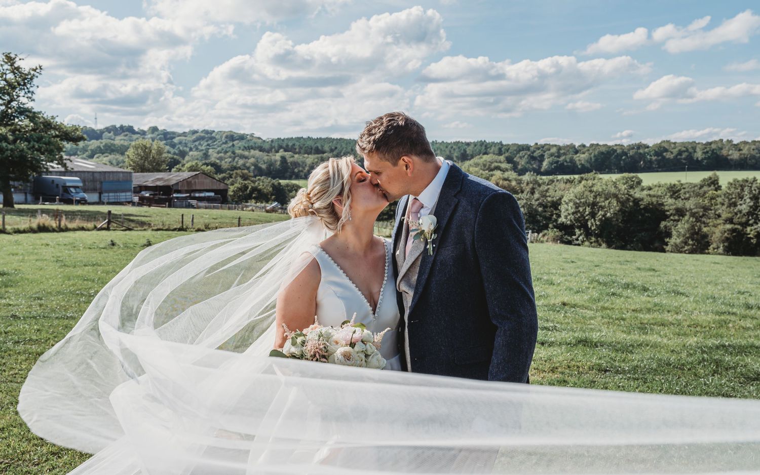 A couple embraces on their wedding day in a scenic countryside setting as the bride's veil flows dramatically in the breeze.