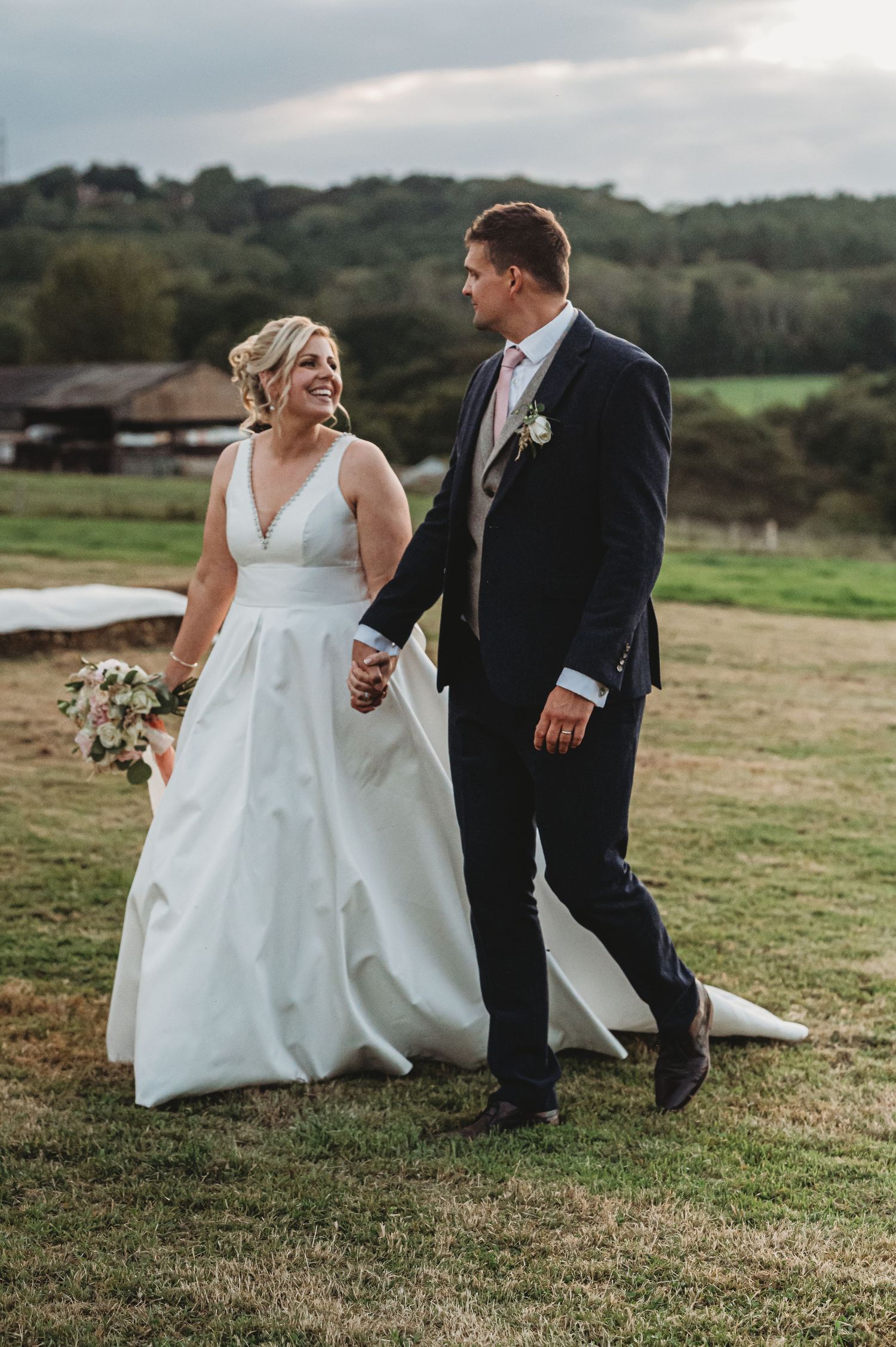 A couple walks hand in hand across a grassy hill during their rustic outdoor wedding ceremony.