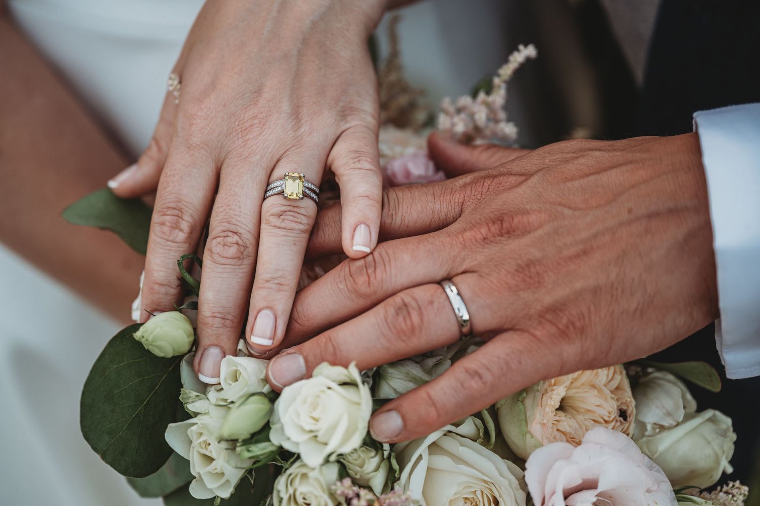A close-up shot of two hands wearing wedding rings resting on a white rose bouquet.