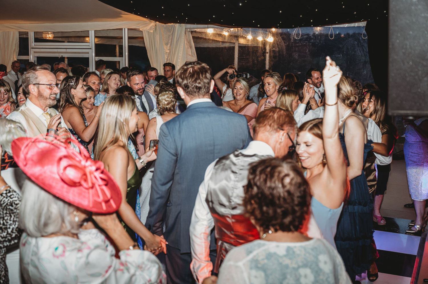 Lively crowd dancing and celebrating at a wedding reception under string lights at night.