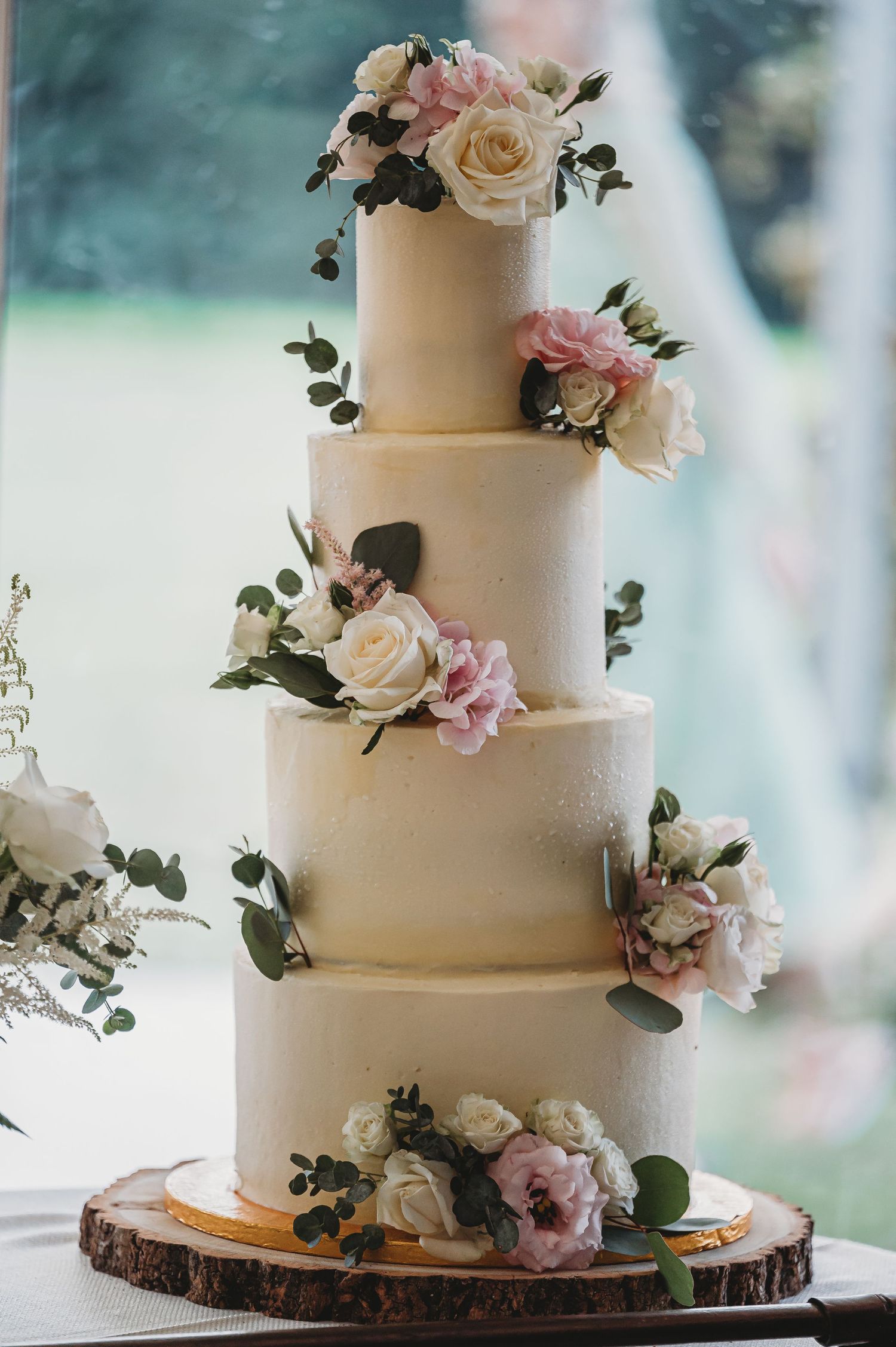 Elegant four-tier white wedding cake decorated with blush pink and cream roses on rustic wood slice base.