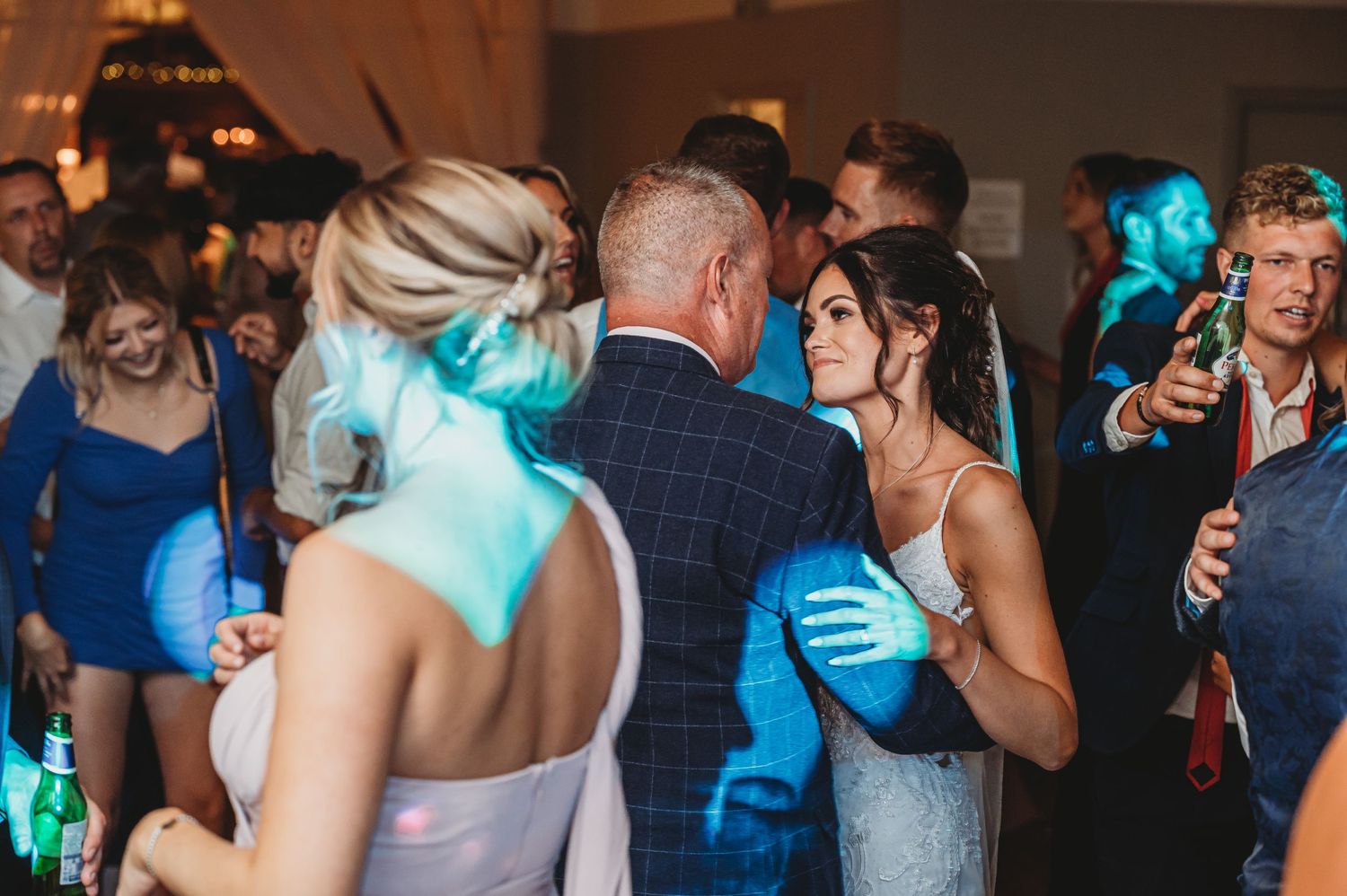 Wedding guests dance under blue lighting at a reception as guests capture the moment on their phones.