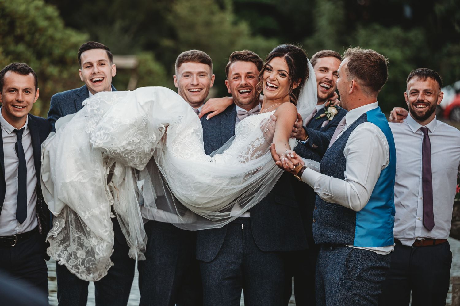 Wedding party groomsmen hold up bride in white dress while laughing together outdoors.