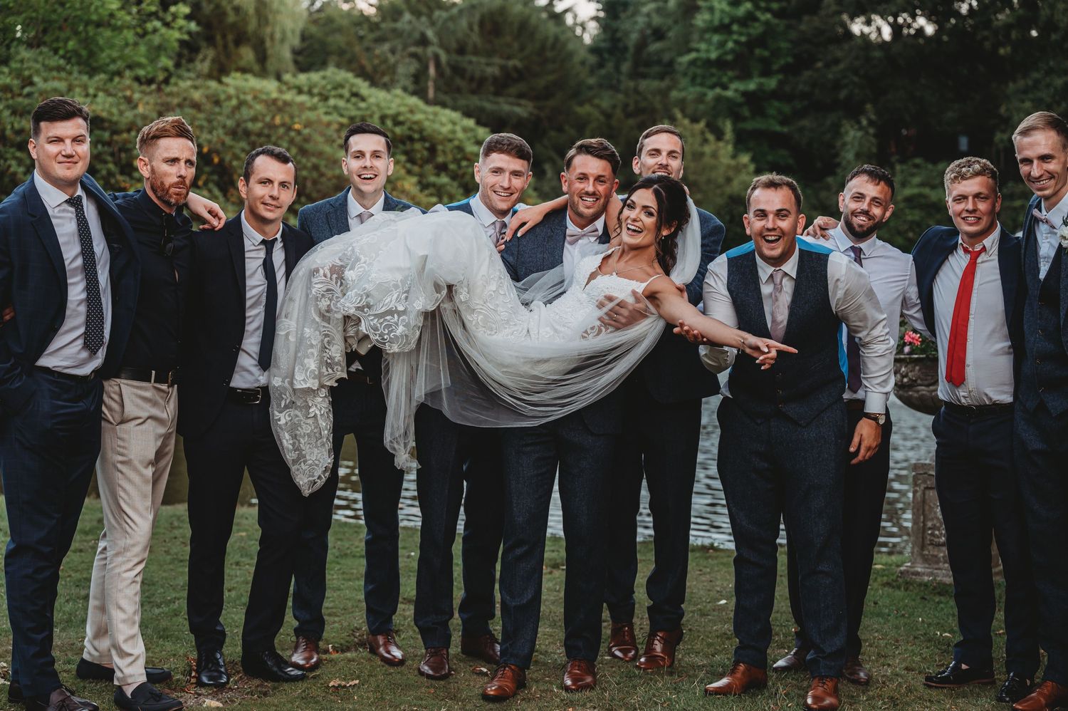 A groomsmen group holds the bride in a white dress in their arms while standing outdoors on a lawn.