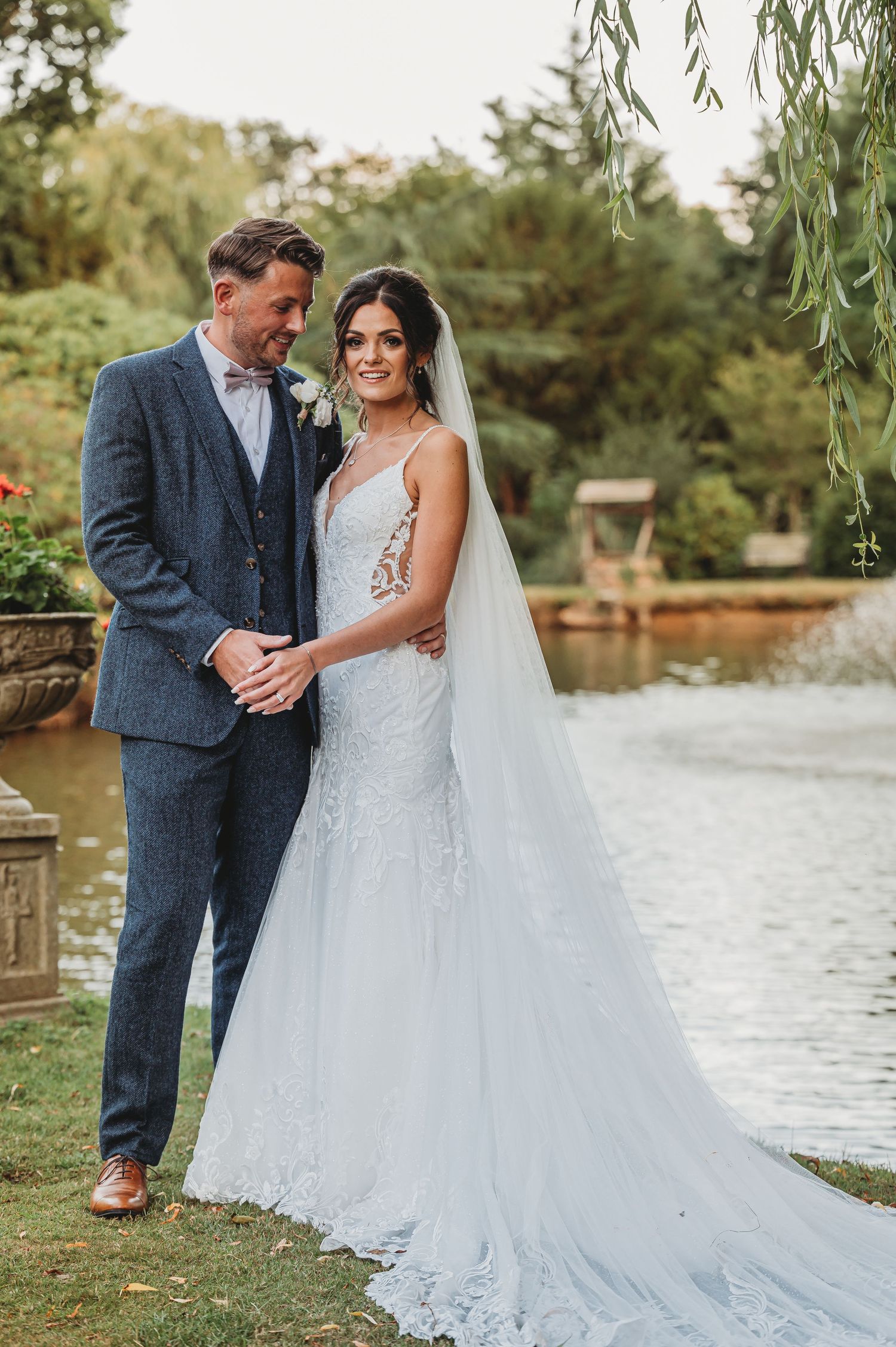 A couple in formal wedding attire poses together by a pond under a willow tree during an outdoor ceremony.