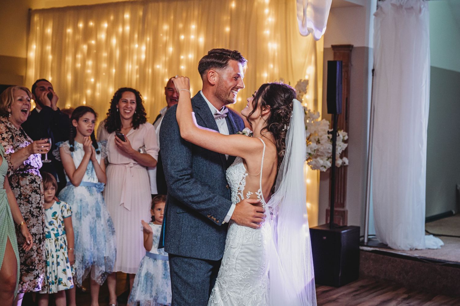 A couple shares their first dance as newlyweds while guests applaud at a wedding reception with twinkle lights in background.