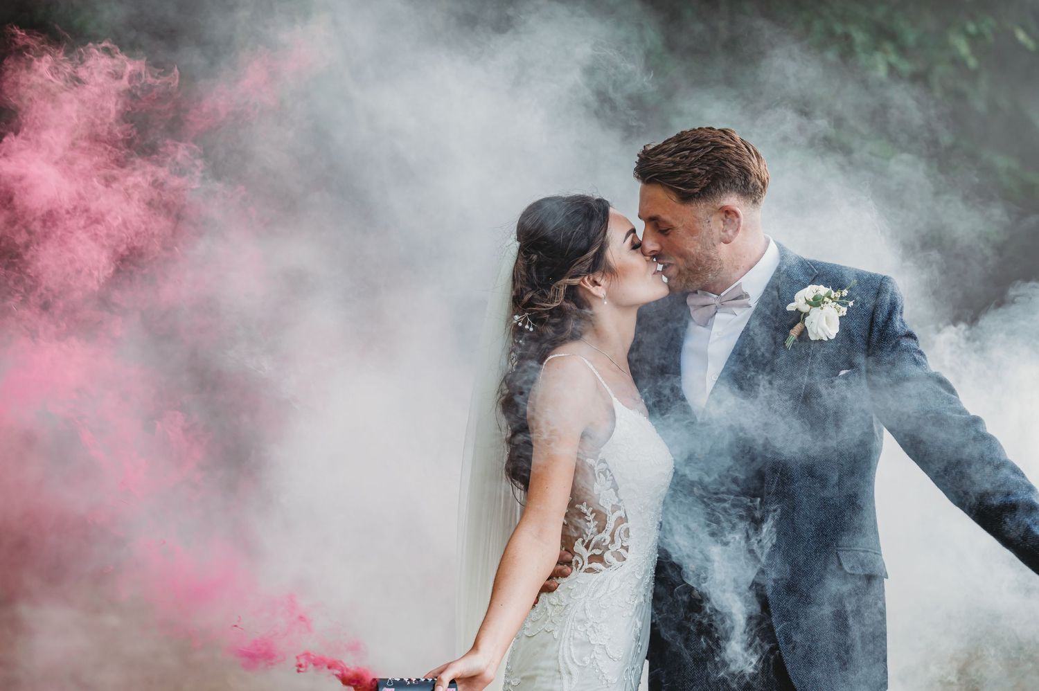 A romantic wedding photo with pink smoke effects and misty atmosphere creating a dreamy outdoor setting.
