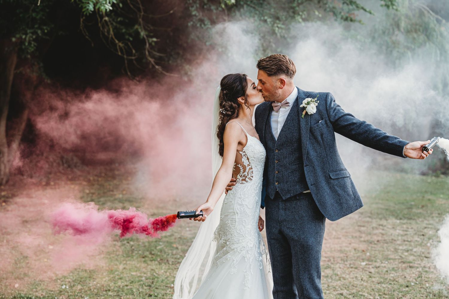 Newlyweds celebrate with pink smoke bombs during outdoor wedding photoshoot in a misty garden setting.