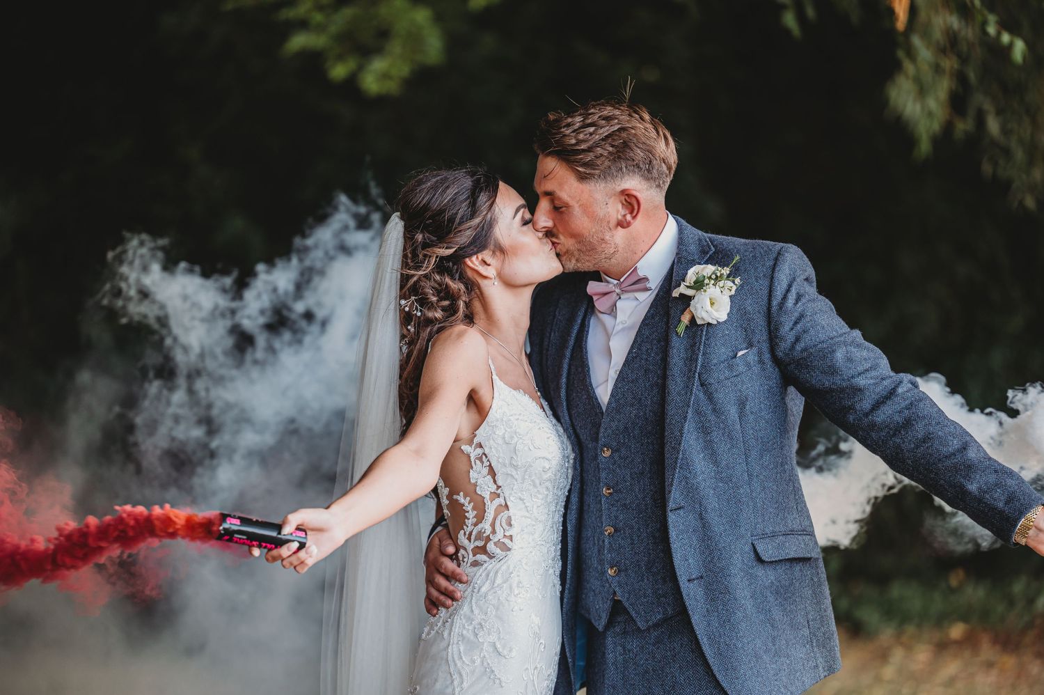 Wedding couple shares romantic moment with red smoke bomb creating dramatic effect in outdoor setting.