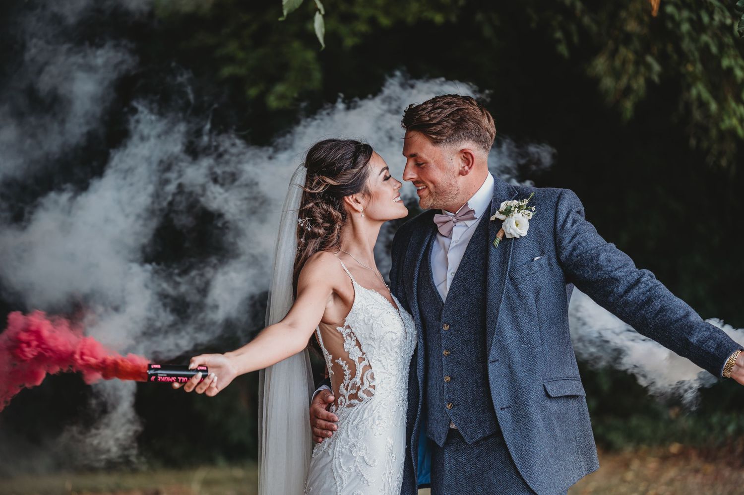 A couple in wedding attire shares a romantic moment amid swirling smoke and red flares in an outdoor setting.