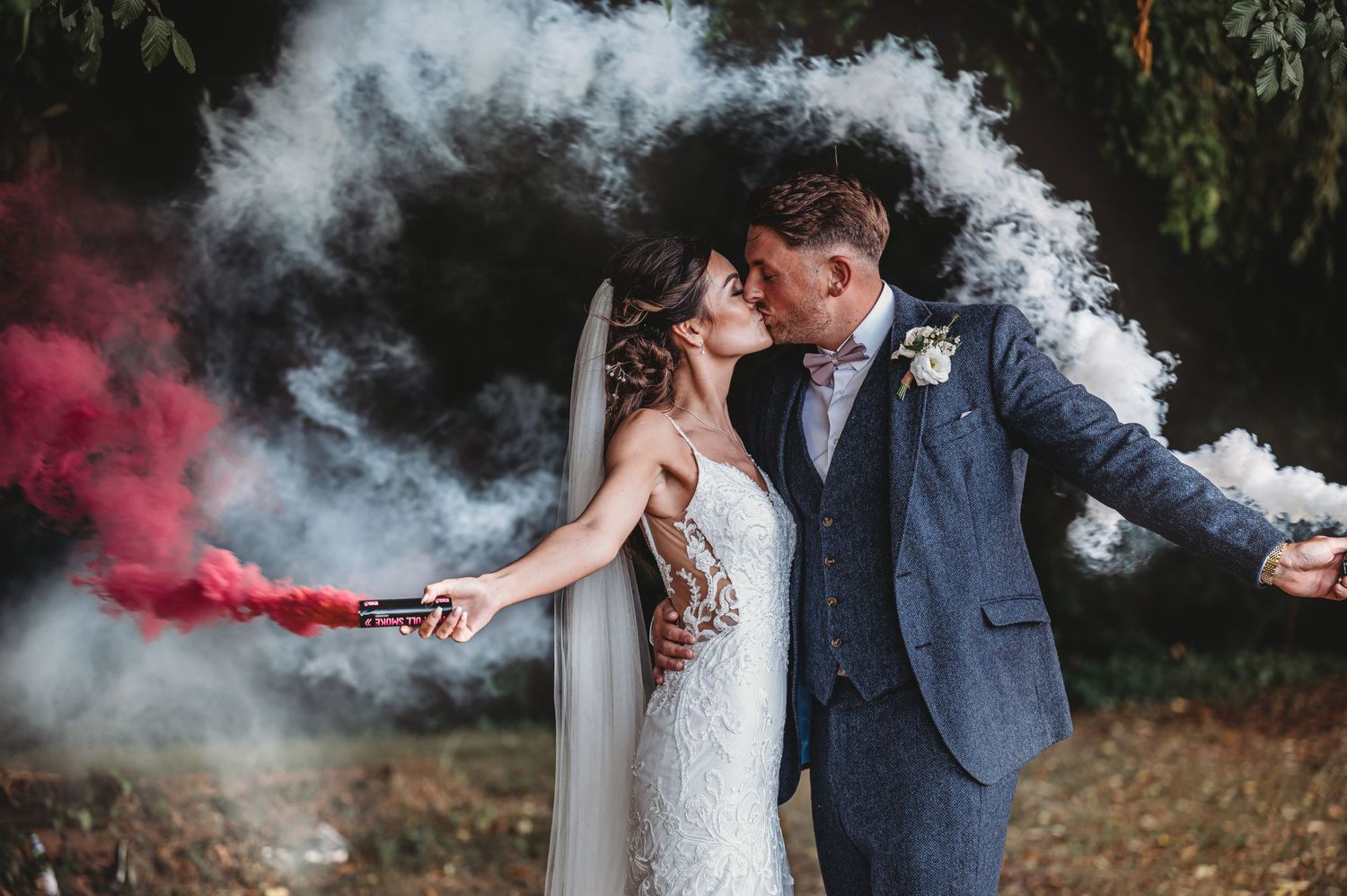 Newlywed couple poses with colorful smoke bombs creating dramatic effect during outdoor wedding photography session.
