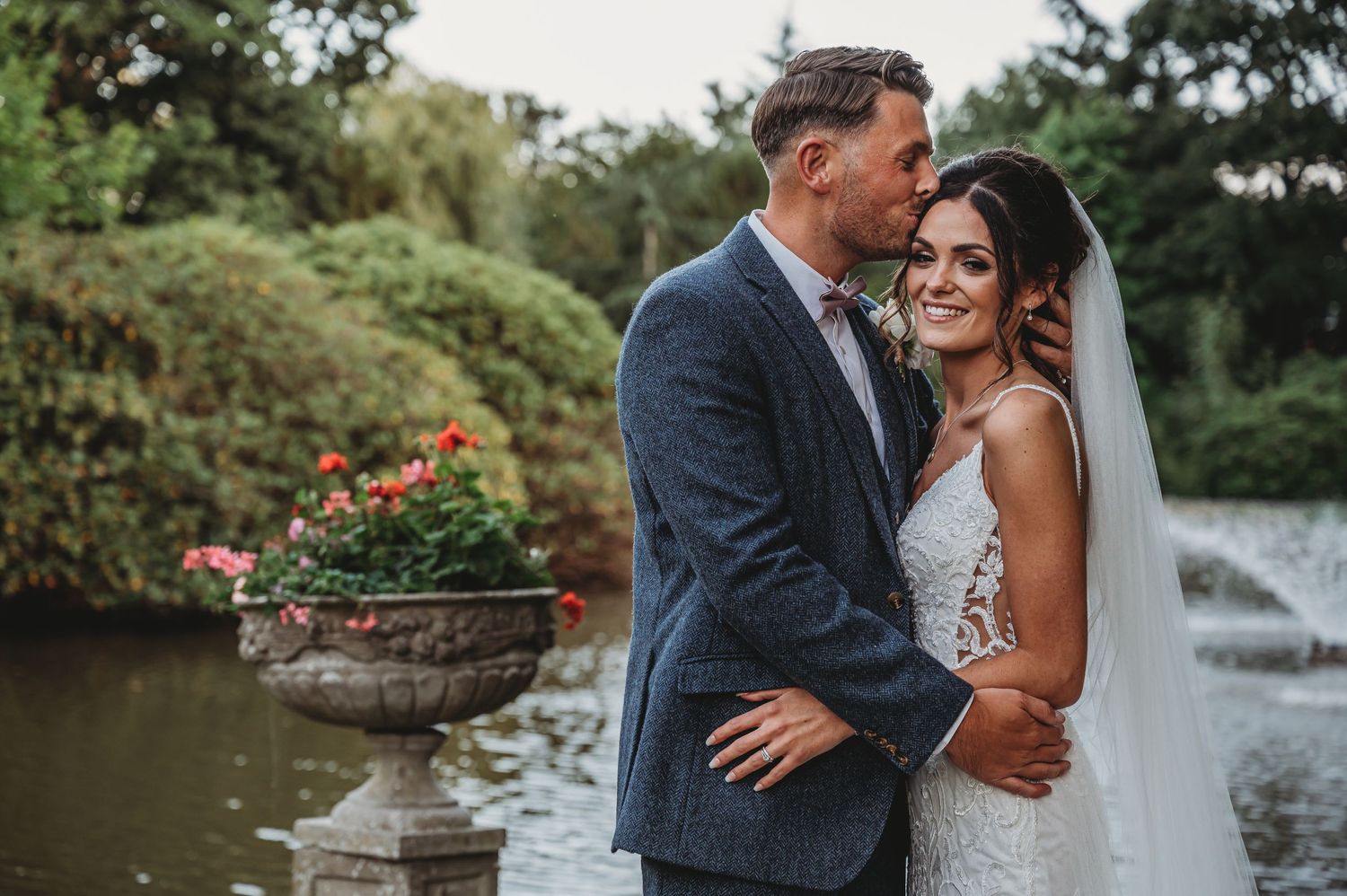 A romantic wedding couple embraces by a garden pond with fountain and flowers in the background.