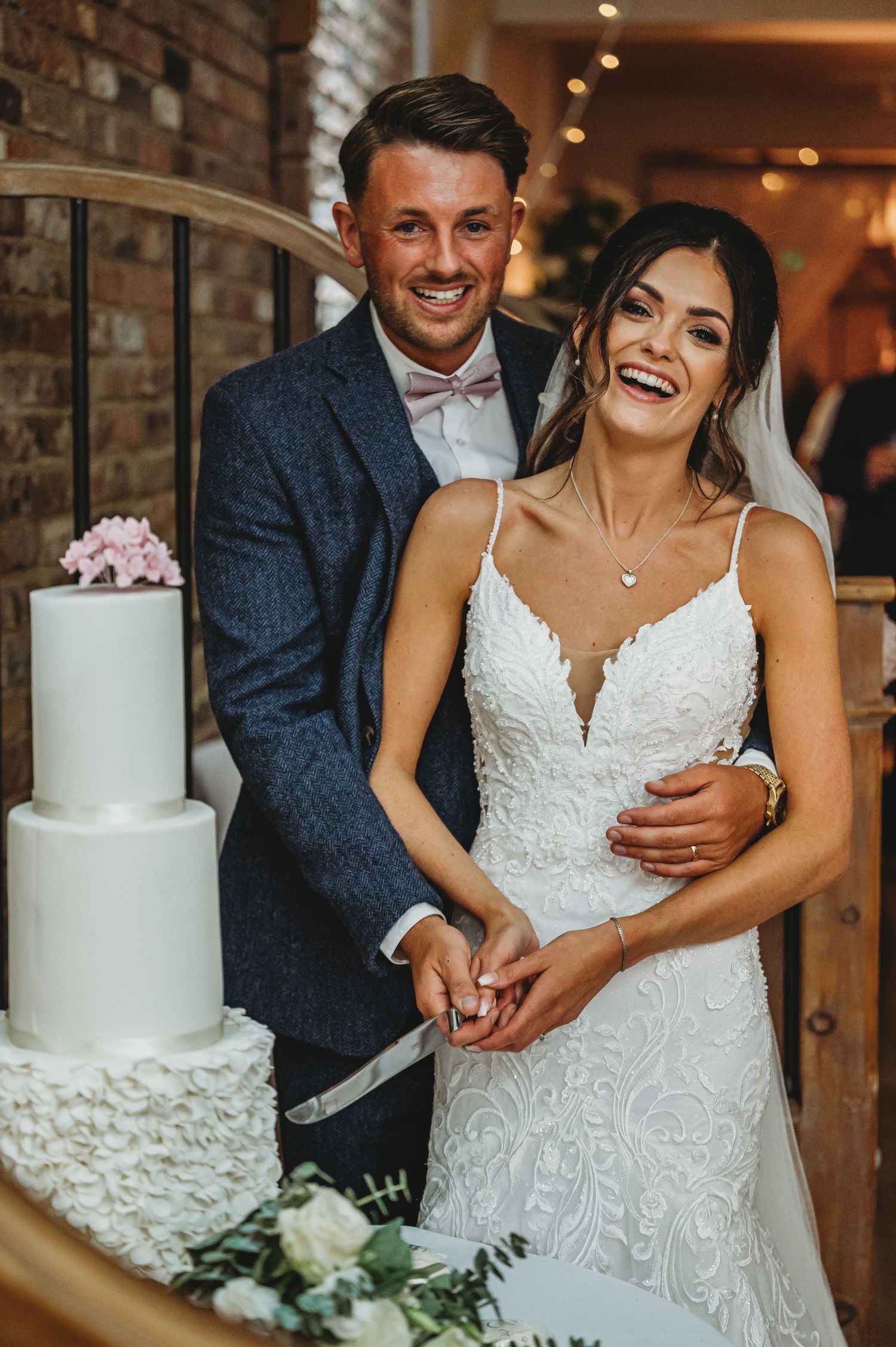 Happy newlyweds share a laugh while cutting their elegant white three-tiered wedding cake at their reception.