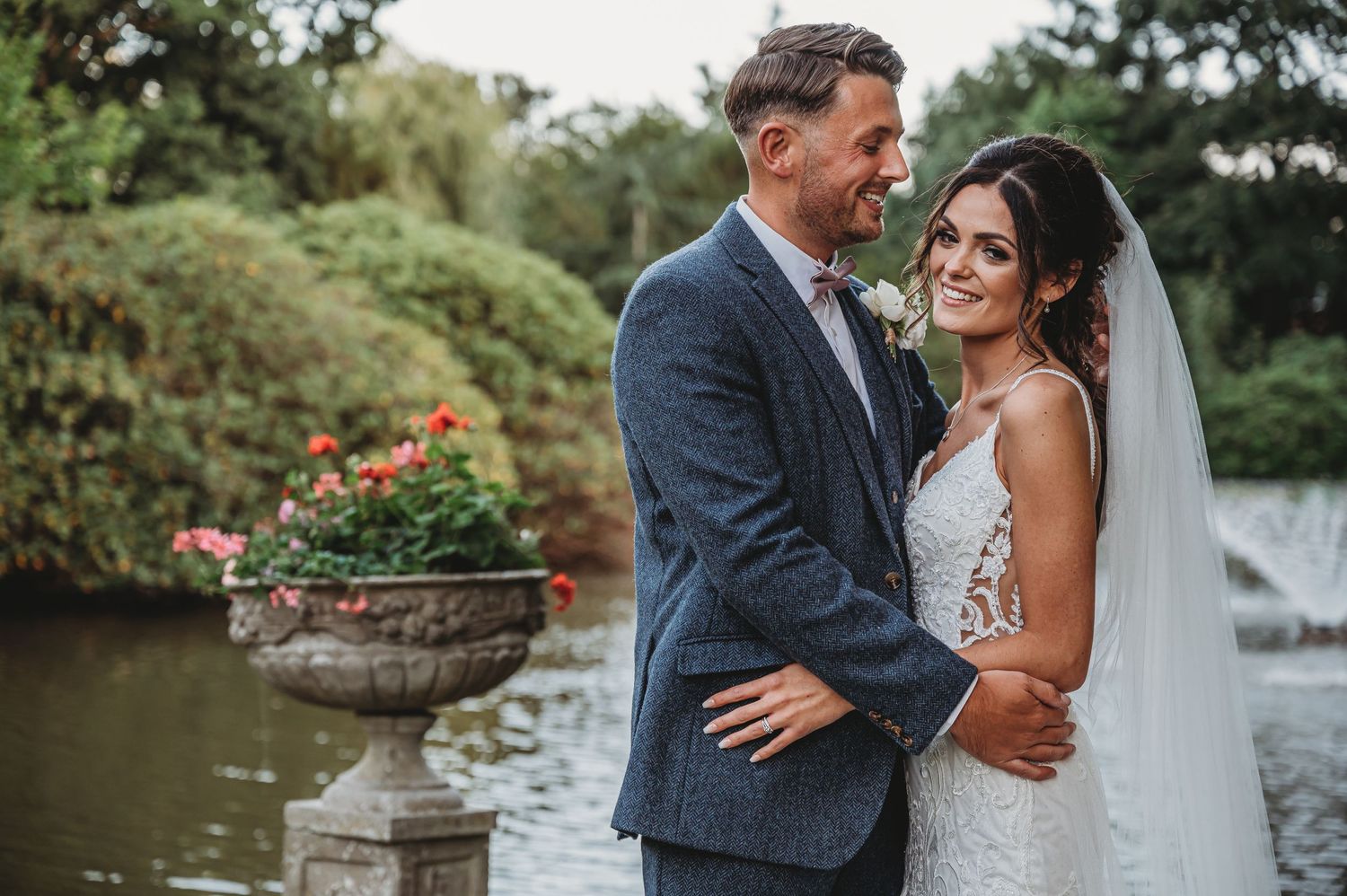A romantic wedding photograph of a couple embracing in a garden with an ornate stone urn and flowering plants.